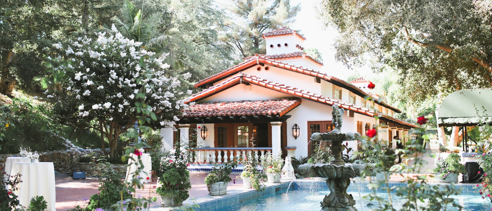 A Spanish-style house with a red-tiled roof situated in a lush garden with blooming plants and trees, including a white flowering tree. A fountain with water features and decorative stone sculptures is in the foreground, and patio furniture is visible on a brick-paved patio.