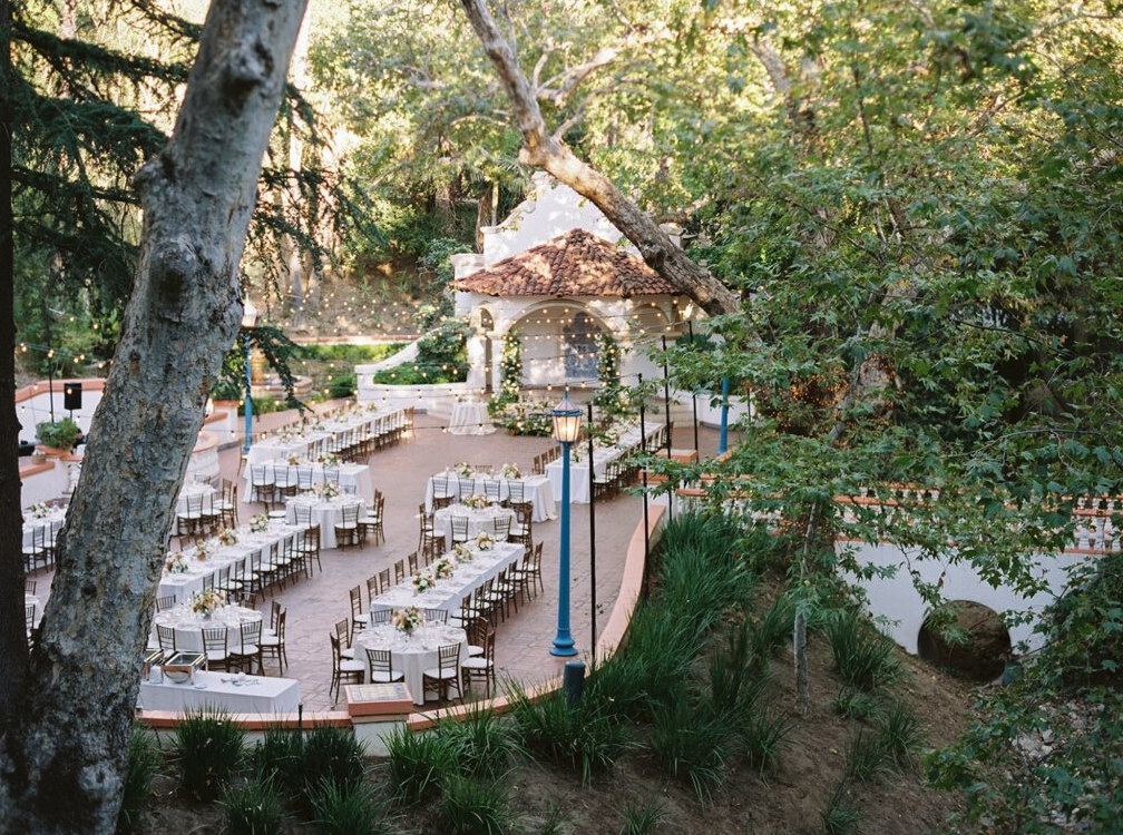 Outdoor wedding reception setup in a lush garden with multiple tables, chairs, floral arrangements, string lights, and a small building with a tiled roof in the background.