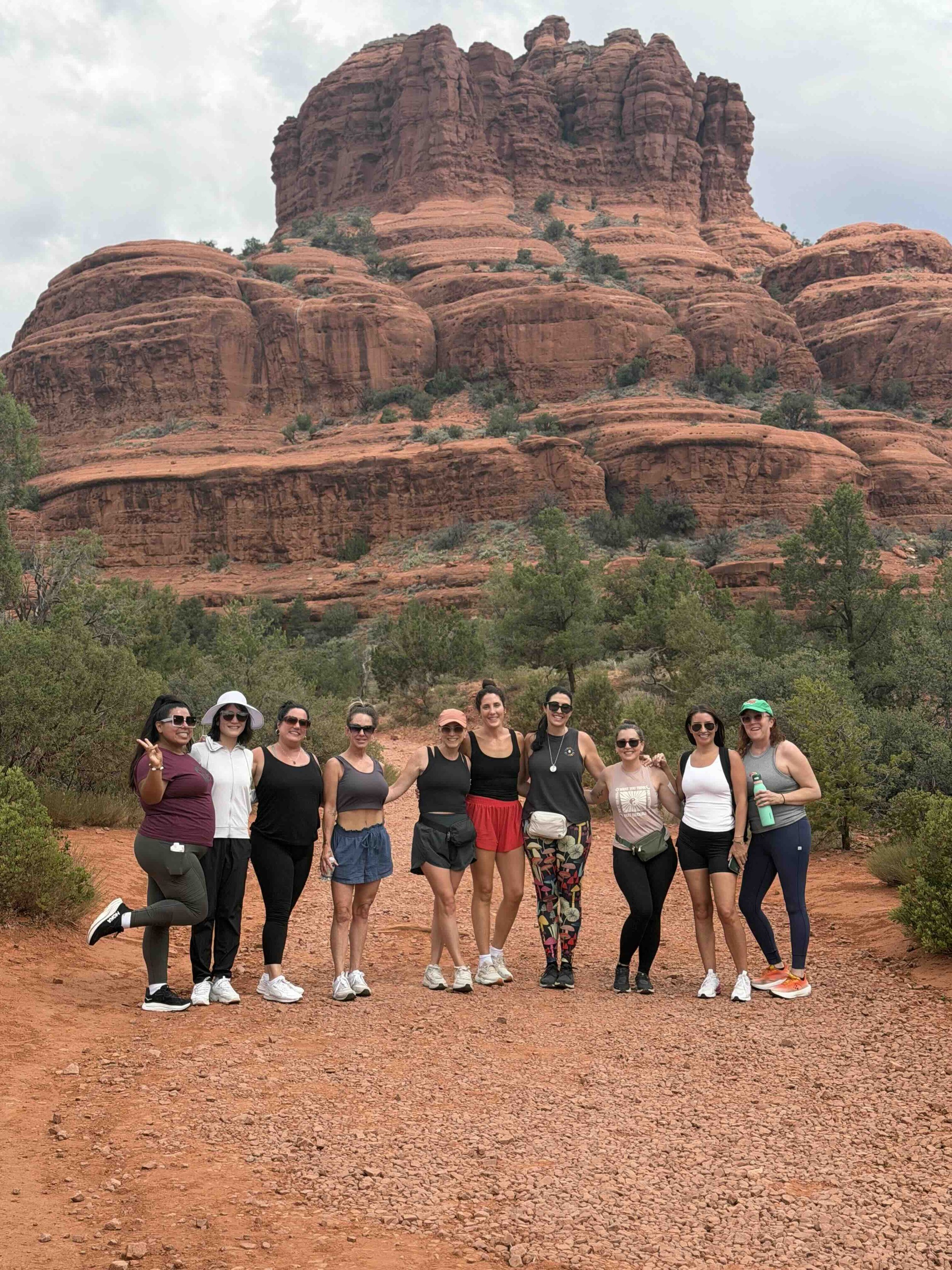 A group of ten women posing together outdoors in front of a large red rock formation in a desert landscape, with trees and cloudy sky in the background.