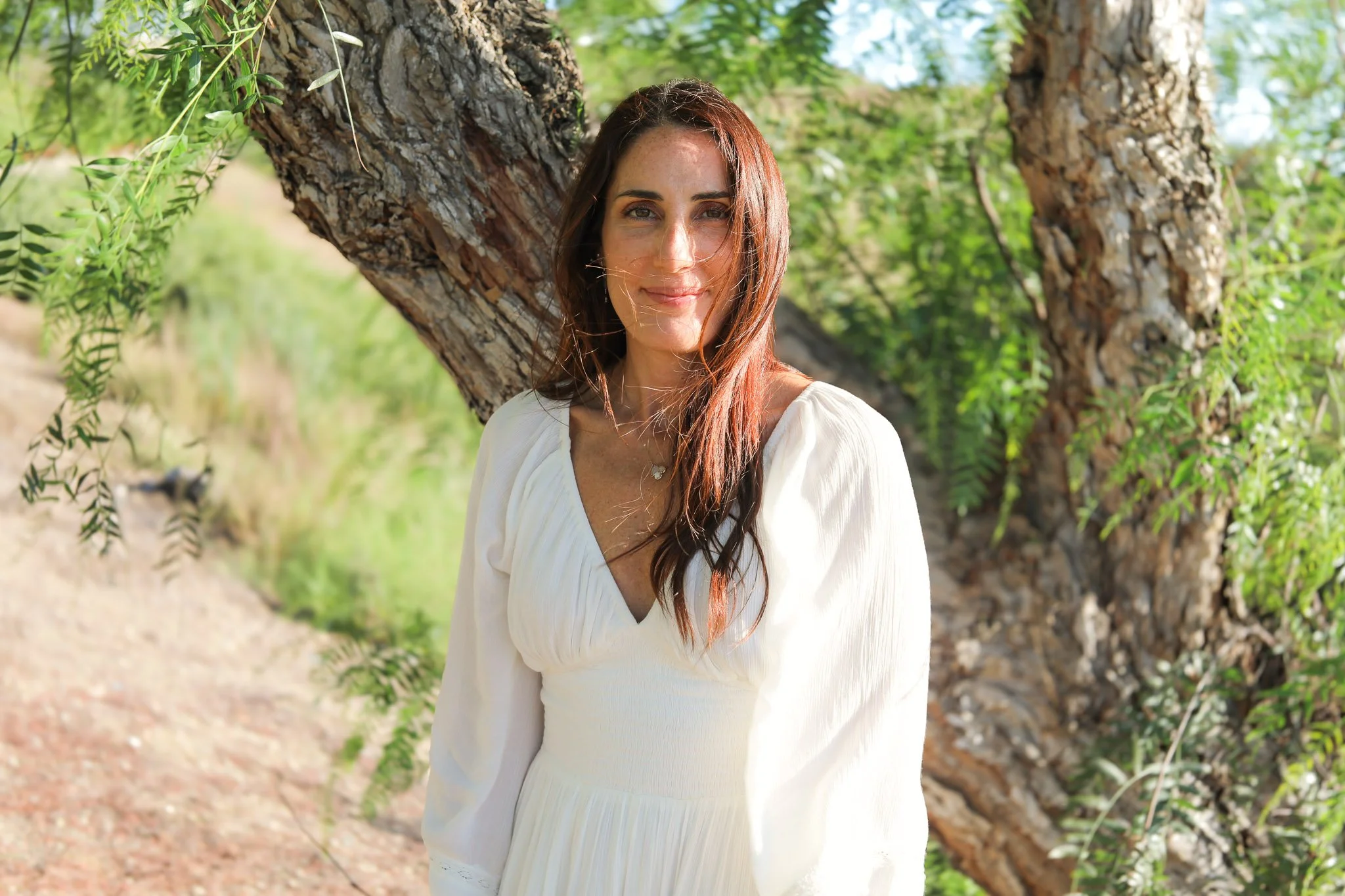A woman with long brown hair and light skin is standing outdoors next to a large tree with green leaves. She is wearing a white blouse and smiling softly at the camera. The background features green foliage and a dirt path.