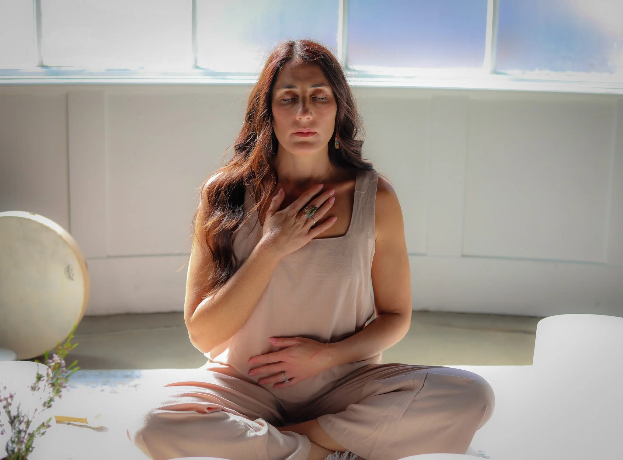 A woman practicing meditation, sitting cross-legged on a mat near a large window with a bright sky outside, her hands resting on her chest and belly, eyes closed, in a calming, peaceful environment.