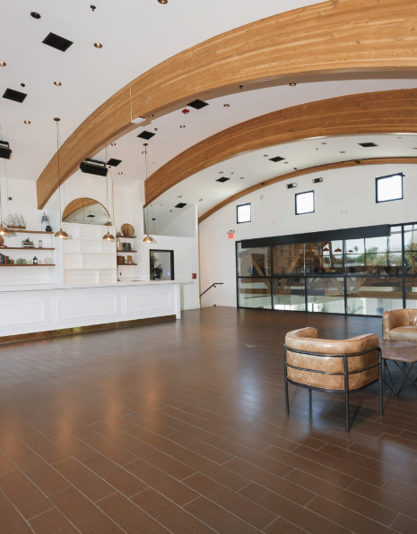 Interior of a modern, spacious room with wooden arched ceiling beams, large windows, a white reception counter, and two tan leather armchairs.