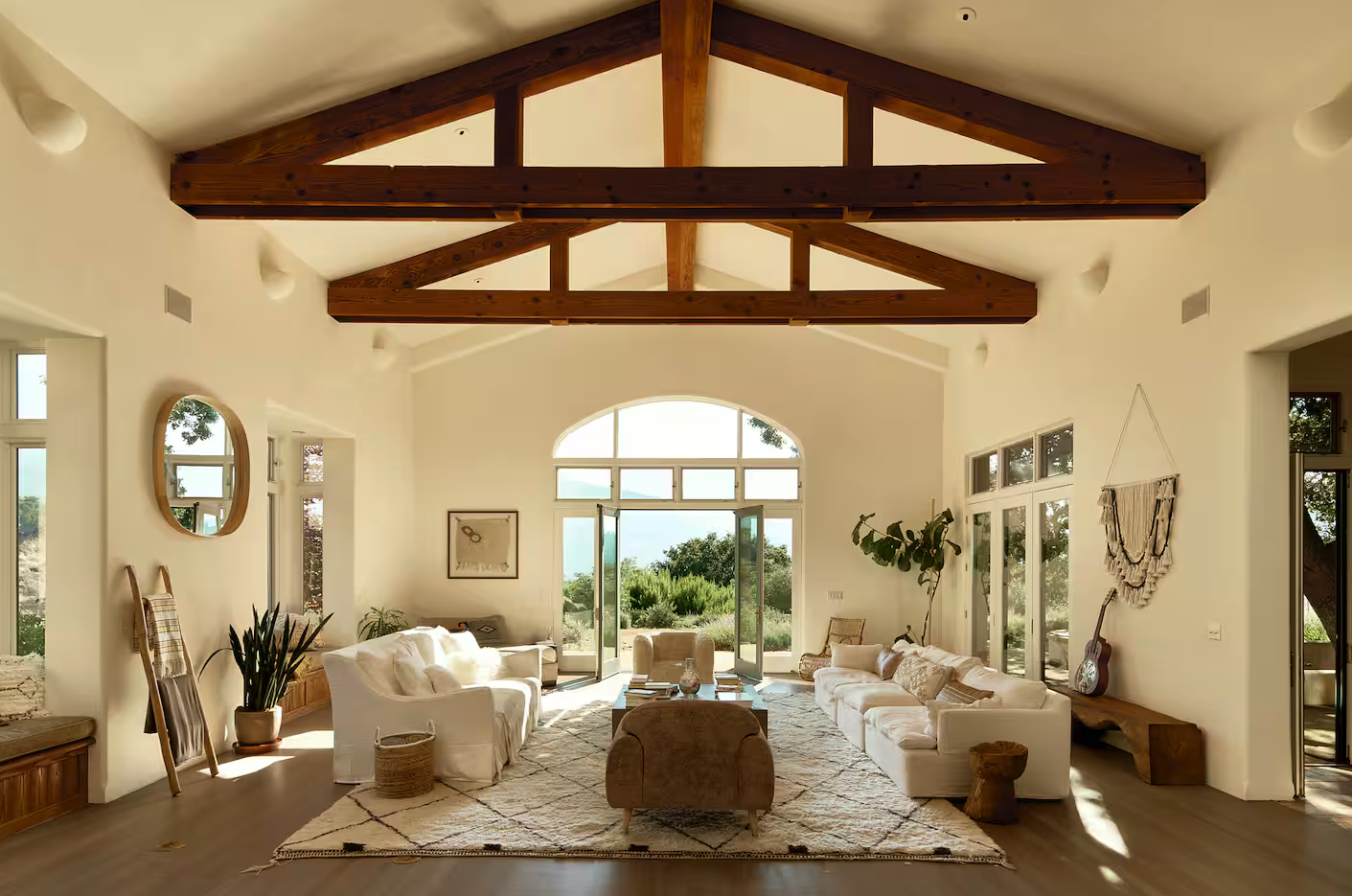 Spacious living room with high vaulted ceiling and exposed wooden beams, large windows and glass doors overlooking greenery, white sofas, plush chairs, and decorative items.
