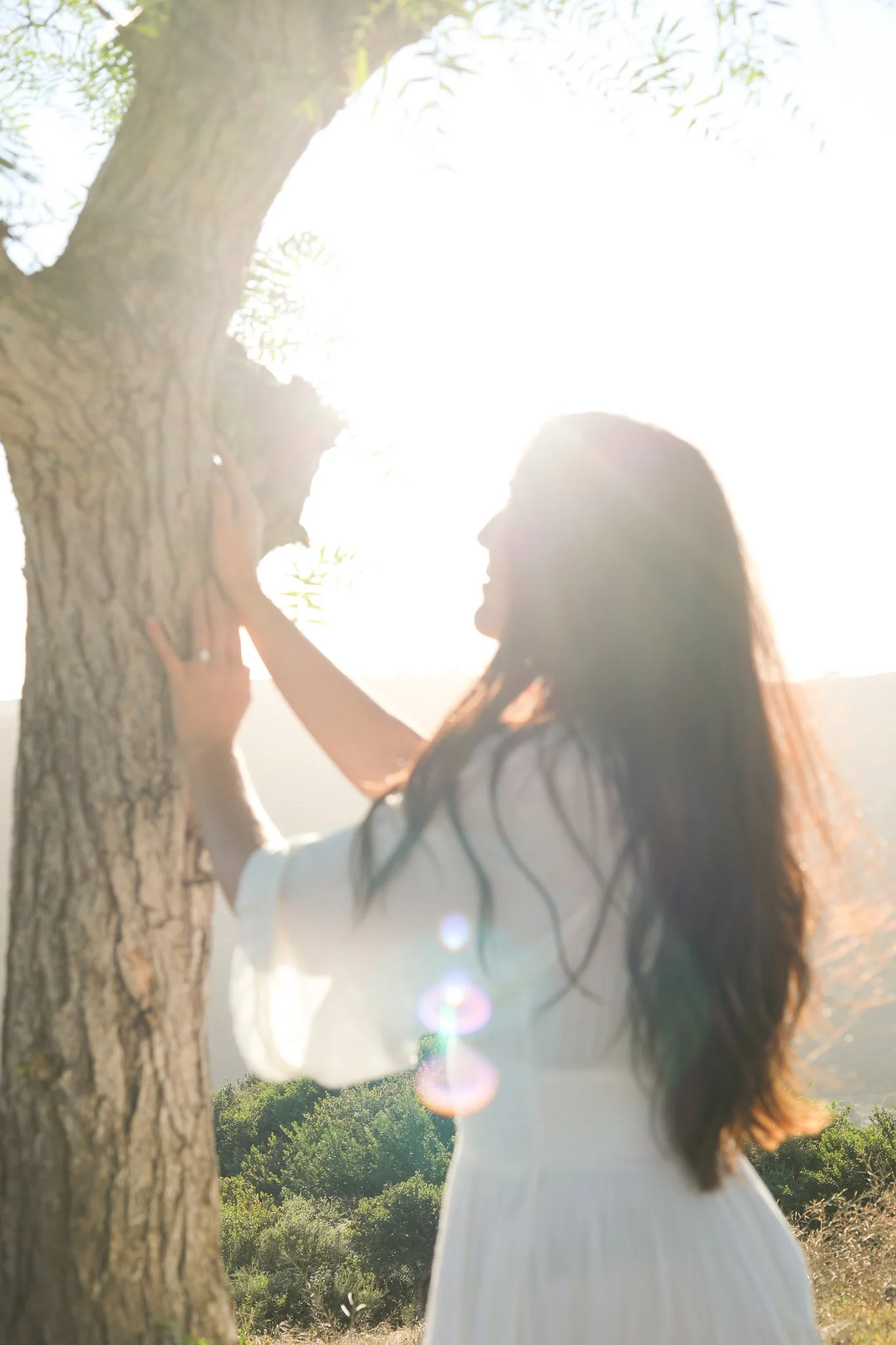A woman with long, dark, wavy hair touching a tree in a sunlit outdoor setting with greenery.