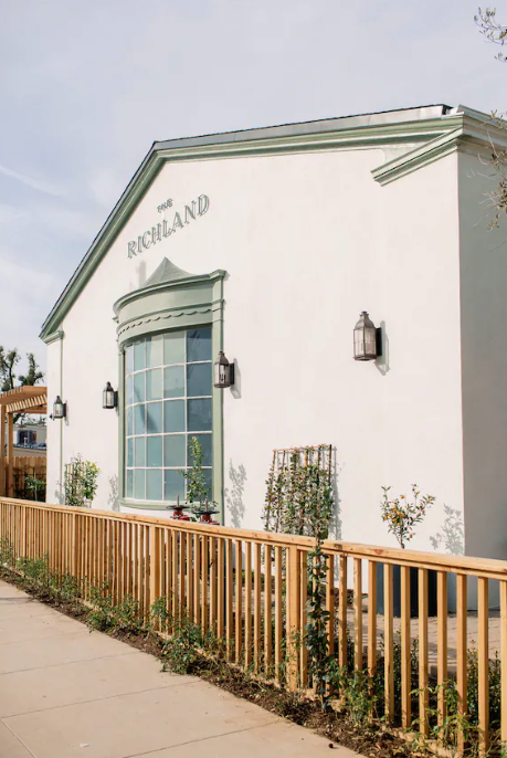 A white building with the word 'Richland' on the front, featuring a large curved window, three black wall-mounted lantern lights, and a wooden fence along a sidewalk with some plants and small trees.