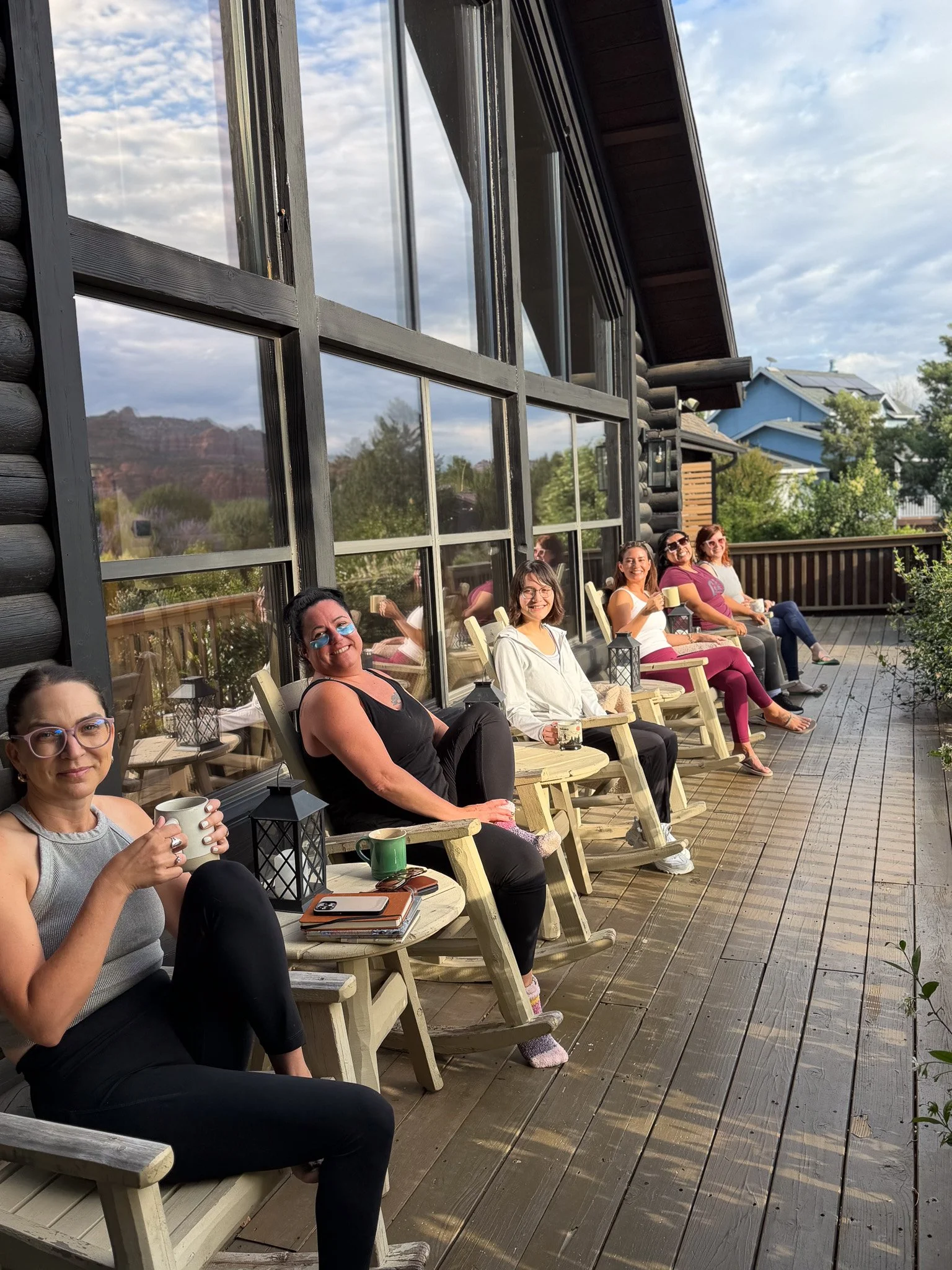 A group of women sitting on a wooden porch, smiling and holding mugs, with mountain scenery and houses in the background.