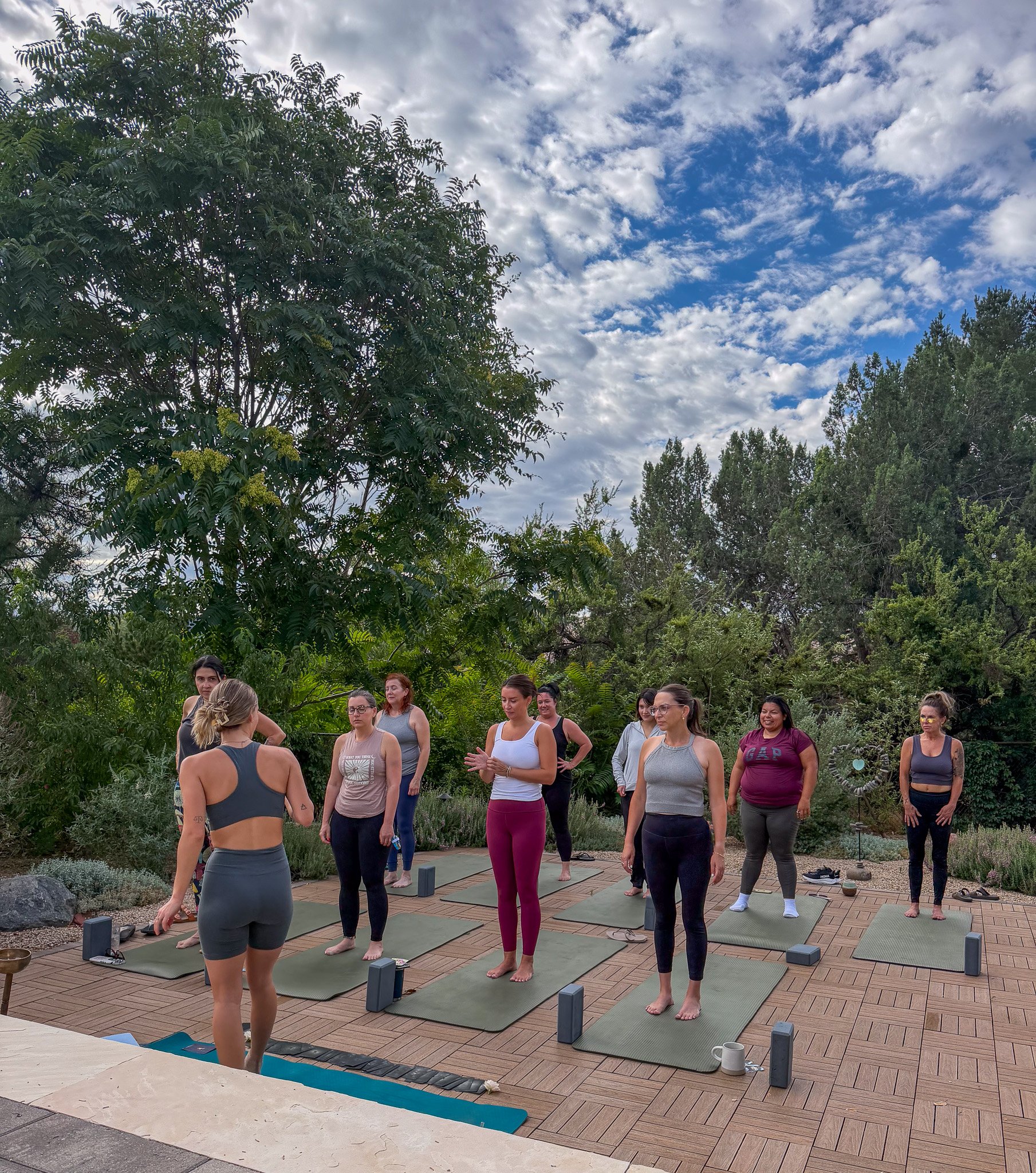 People participating in an outdoor yoga class on mats, surrounded by lush trees and a partly cloudy sky.
