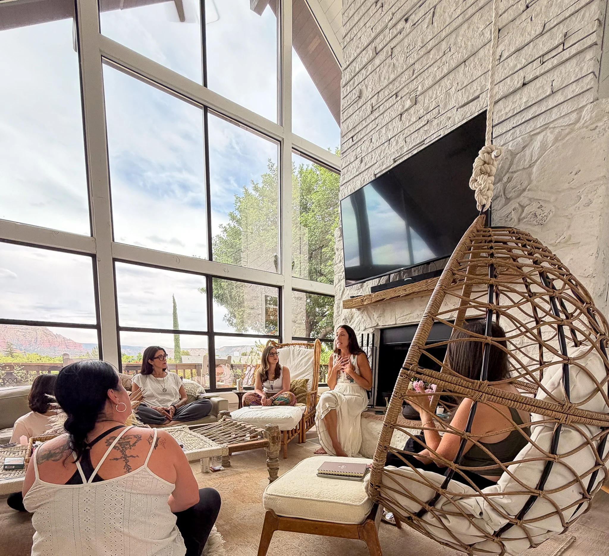 A group of women sitting and talking inside a modern room with large windows, a white stone fireplace, and wicker chairs. The sky is cloudy outside, and natural light fills the space.