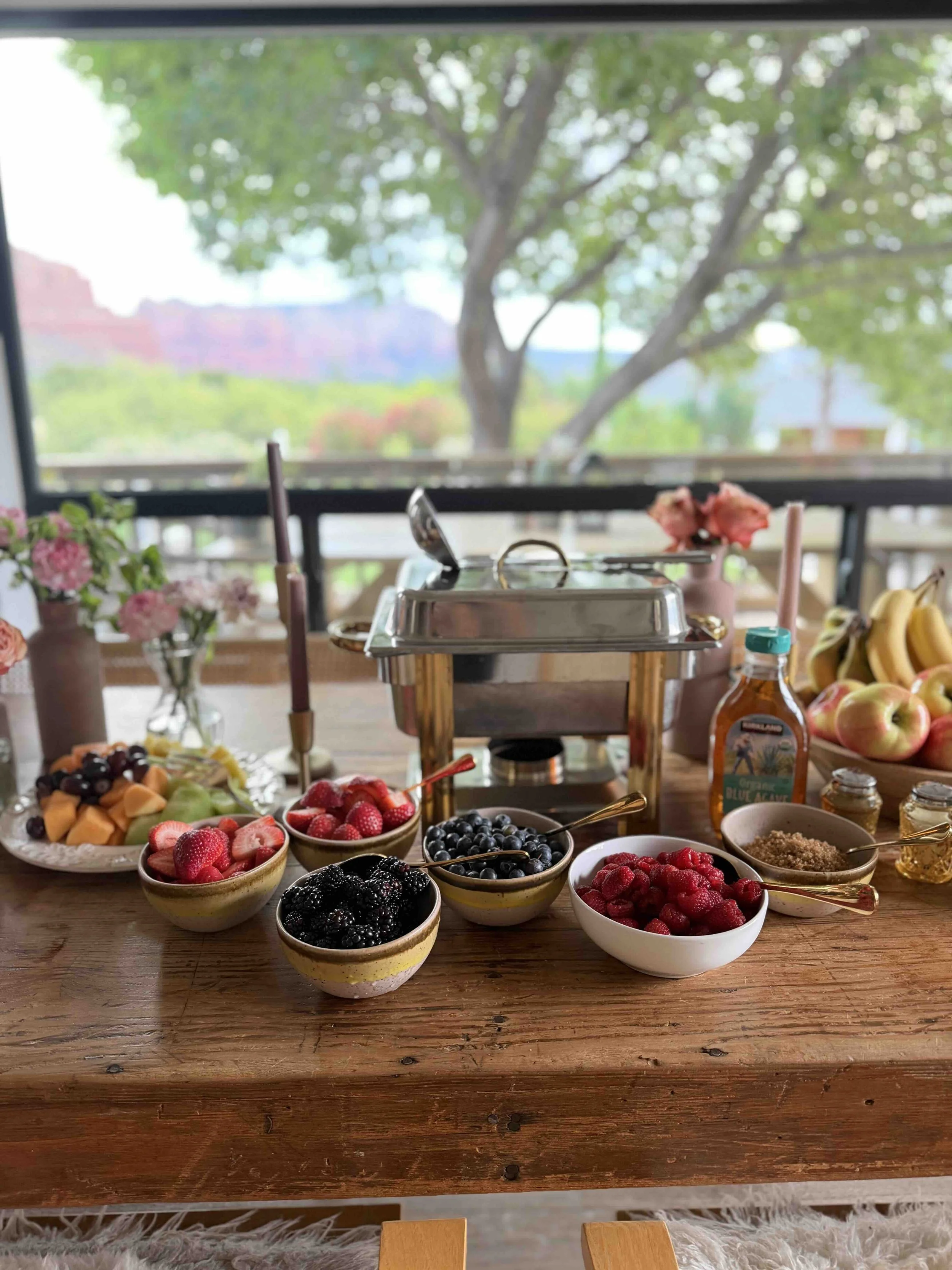 A breakfast spread with bowls of fresh berries, fruit slices, honey, and a chafing dish on a wooden table, with a window showing trees outside.