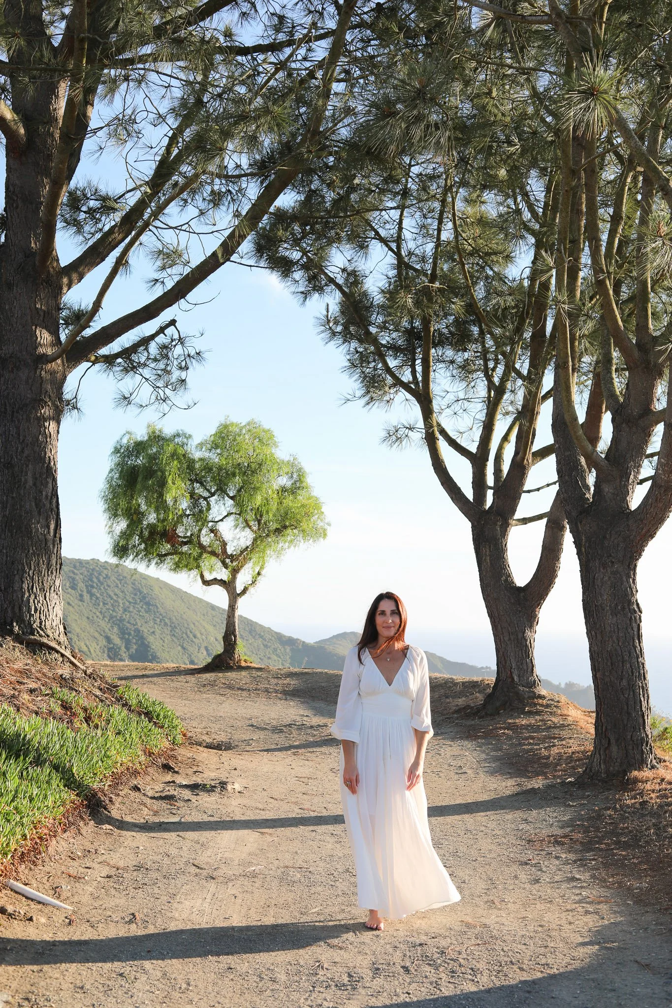 A woman in a long white dress walking barefoot on a dirt path surrounded by trees under a clear sky.