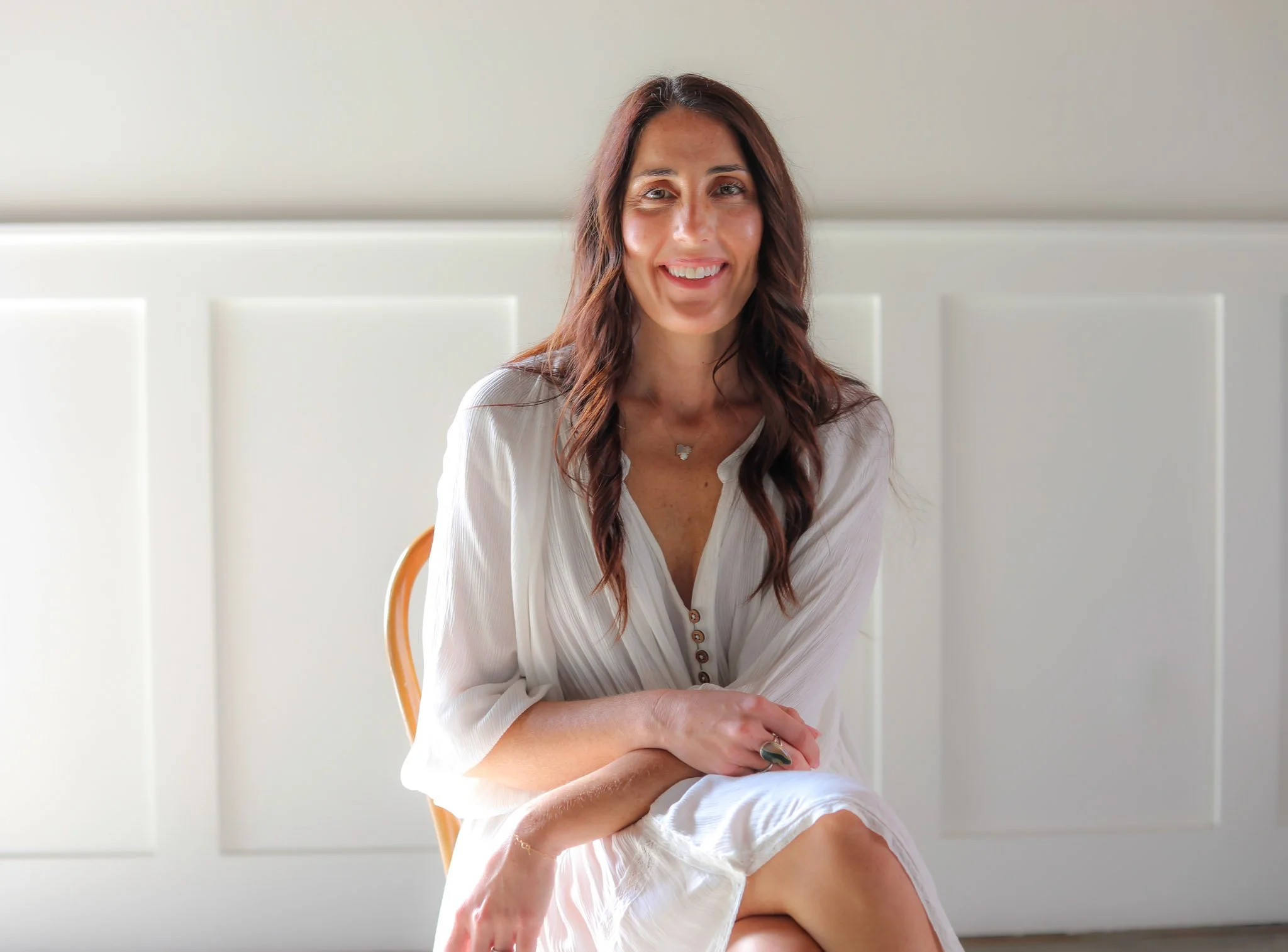 A woman with long reddish-brown hair sitting on a chair, smiling, wearing a white blouse with buttons, and a butterfly shaped necklace, in front of a plain light-colored background.