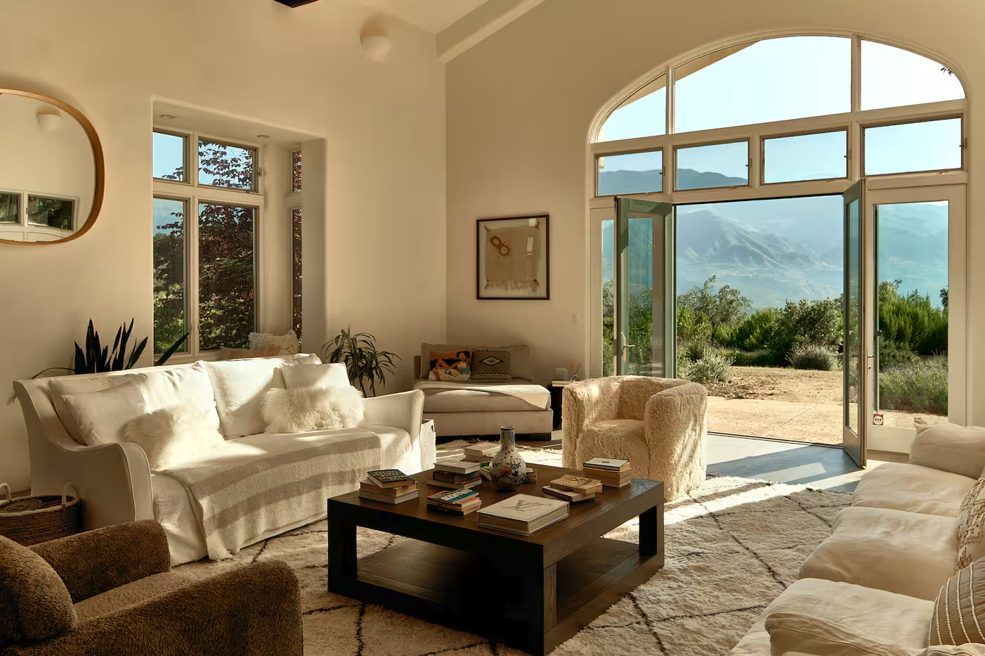 A living room with white sofas, a beige armchair, and a black coffee table with books and decorative items. Large windows and glass doors open to a scenic mountain landscape outside, with natural light flooding the room.