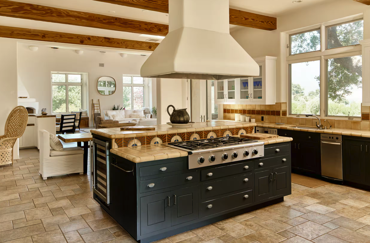 Open-concept kitchen with a large island, black cabinetry, a white range hood, and tiled countertops, leading into a living area with large windows, white walls, and wooden ceiling beams.
