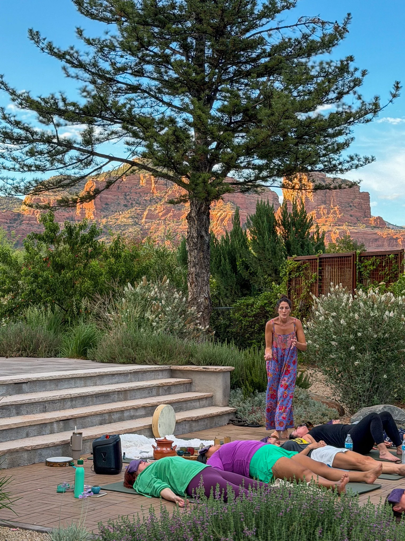 A group of people practicing yoga outdoors on a wooden patio, with some lying on their stomachs, and a woman in colorful clothing standing and instructing. The background includes lush greenery, a large tree, and red rock formations under a blue sky.