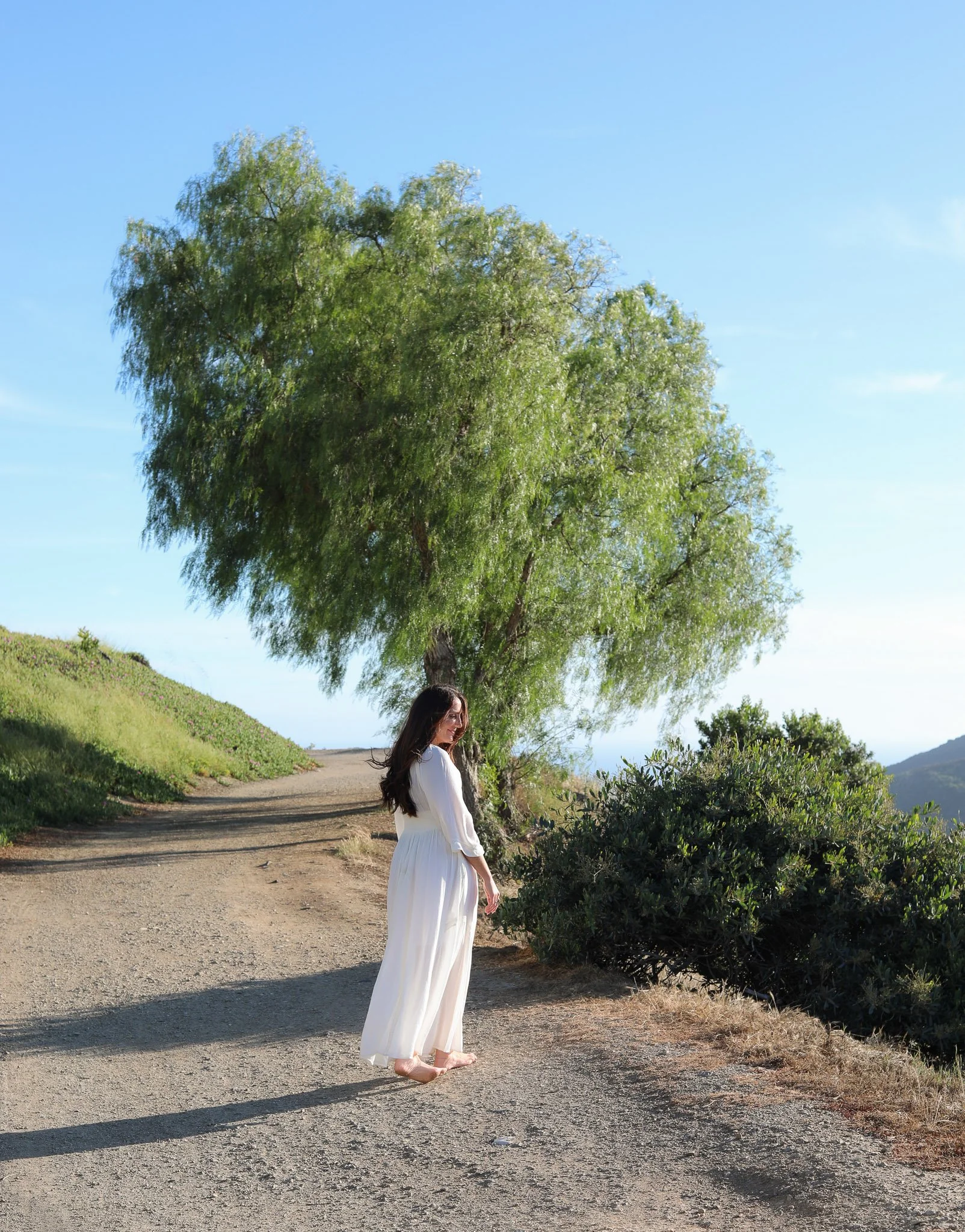 A woman in a white dress walking barefoot on a dirt path in a natural outdoor setting, with a large leafy tree and green bushes around her, under a clear blue sky.