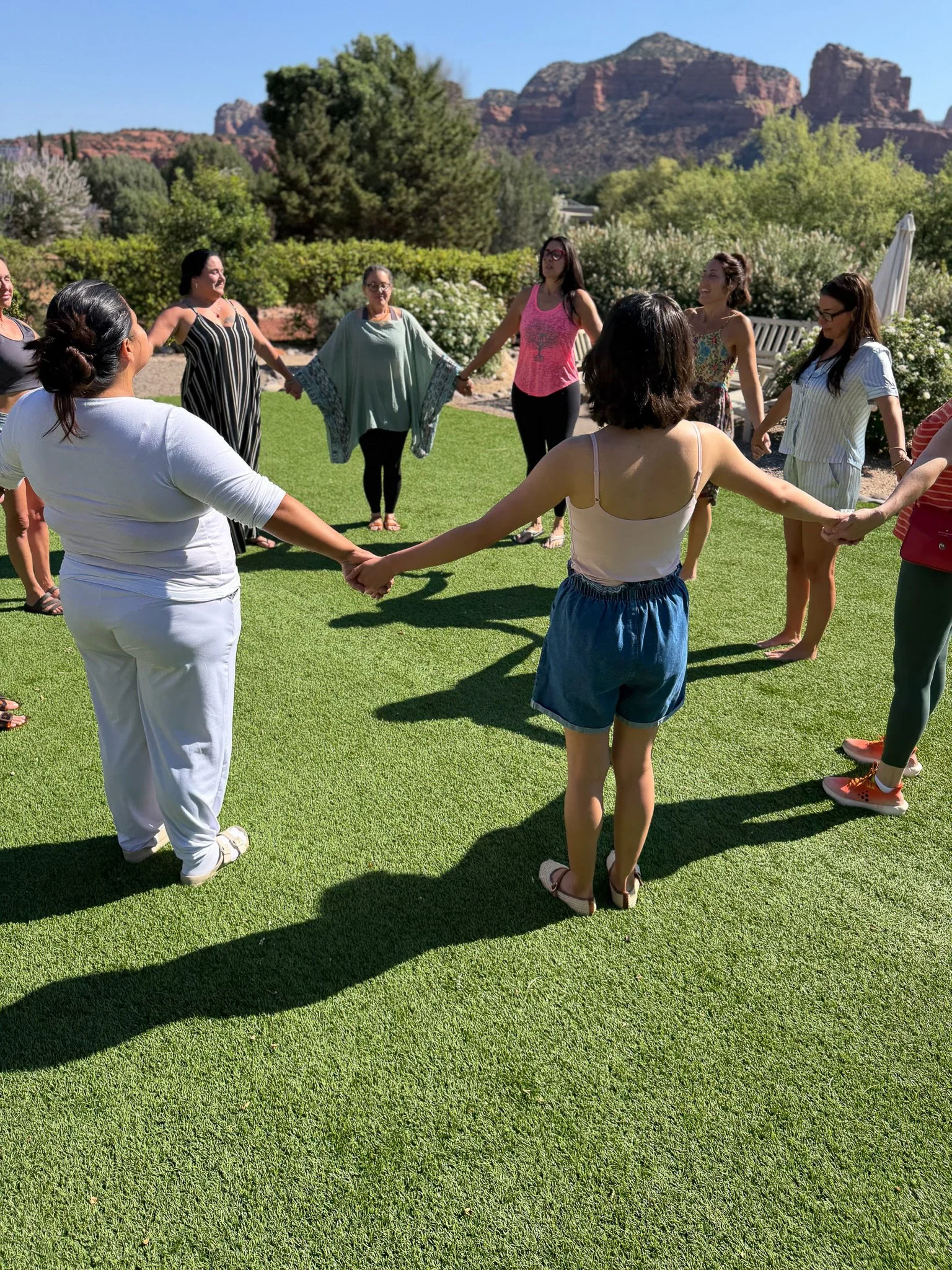 A group of women holding hands in a circle on a grassy field outdoors with trees and mountains in the background.