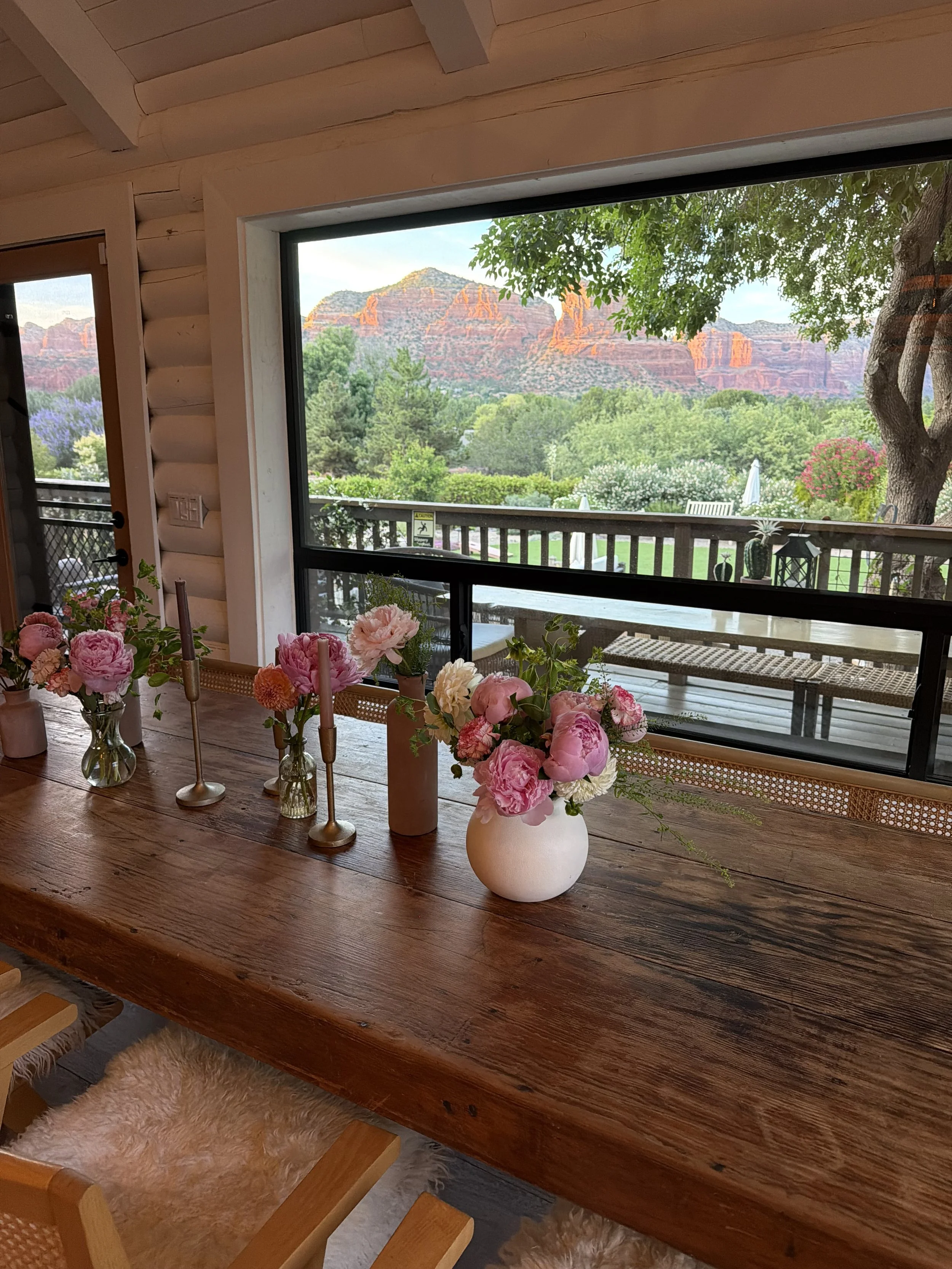 View of a wooden dining table decorated with multiple vases and candles, with a large window showing a scenic landscape of green trees, bushes with flowers, and red rock mountains in the background, along with a balcony with outdoor chairs.