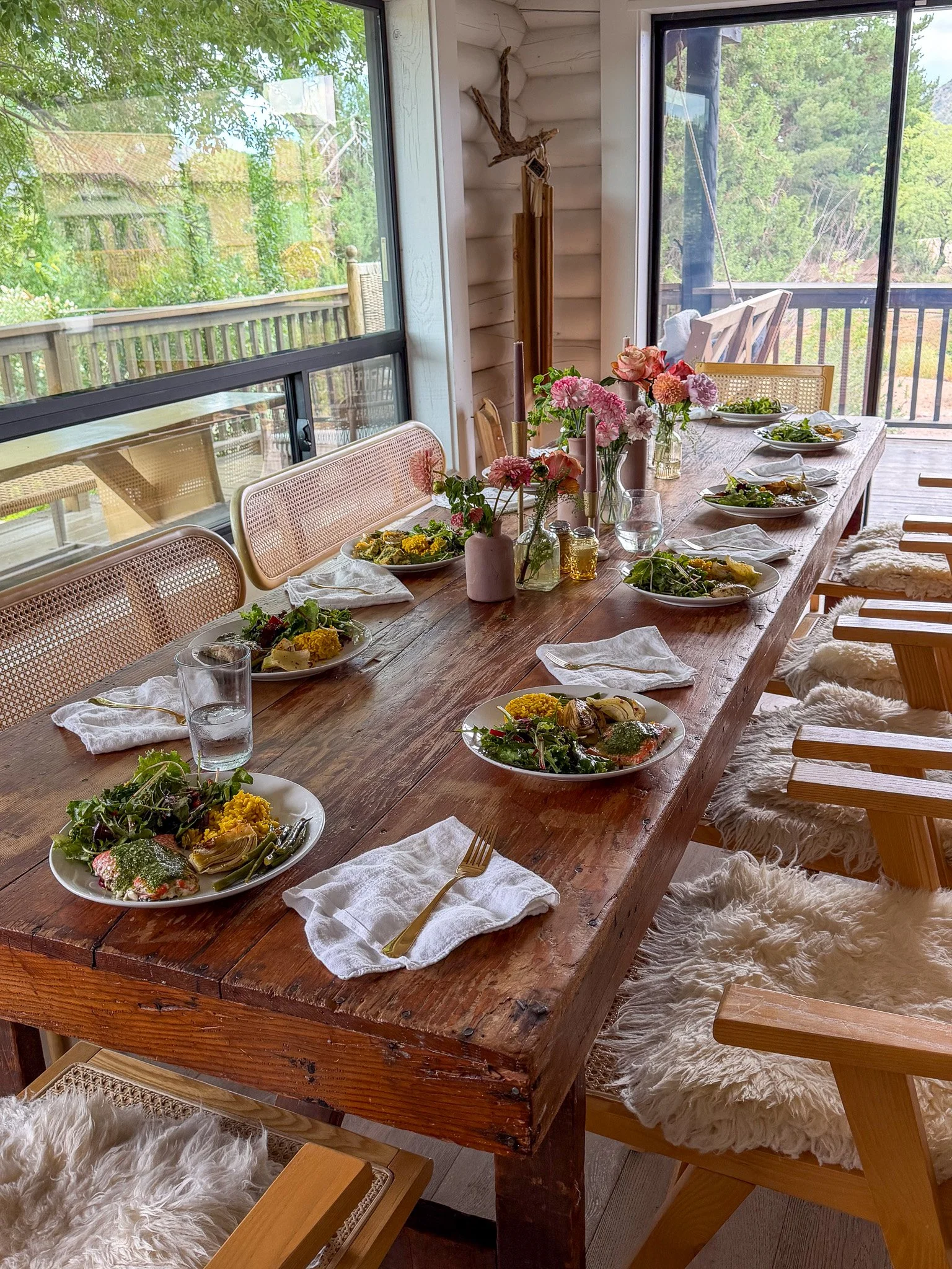 A wooden dining table set for a meal, with plates of salads, glass of water, cutlery on white napkins, pink and orange flowers in vases, candles, and surrounded by wooden chairs with fluffy sheepskin cushions, in a bright room with large windows showing green trees outside.