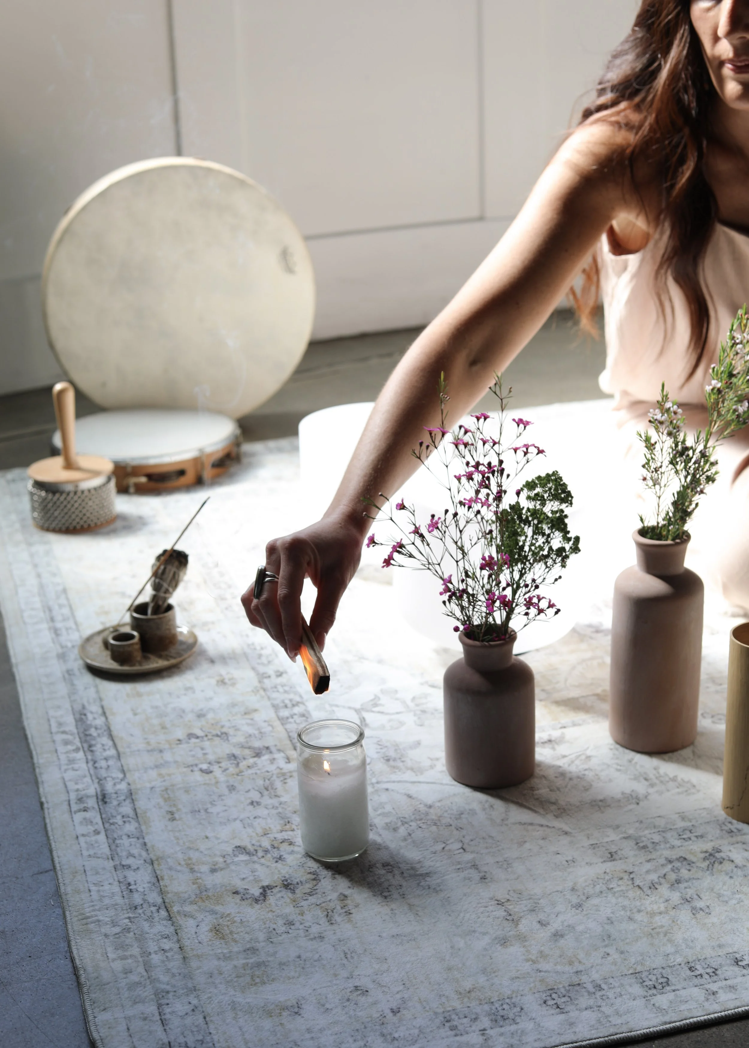 A woman arranging pink and purple flowers in pink and beige vases on a patterned tablecloth, with candles, a drum, and gardening tools in the background.