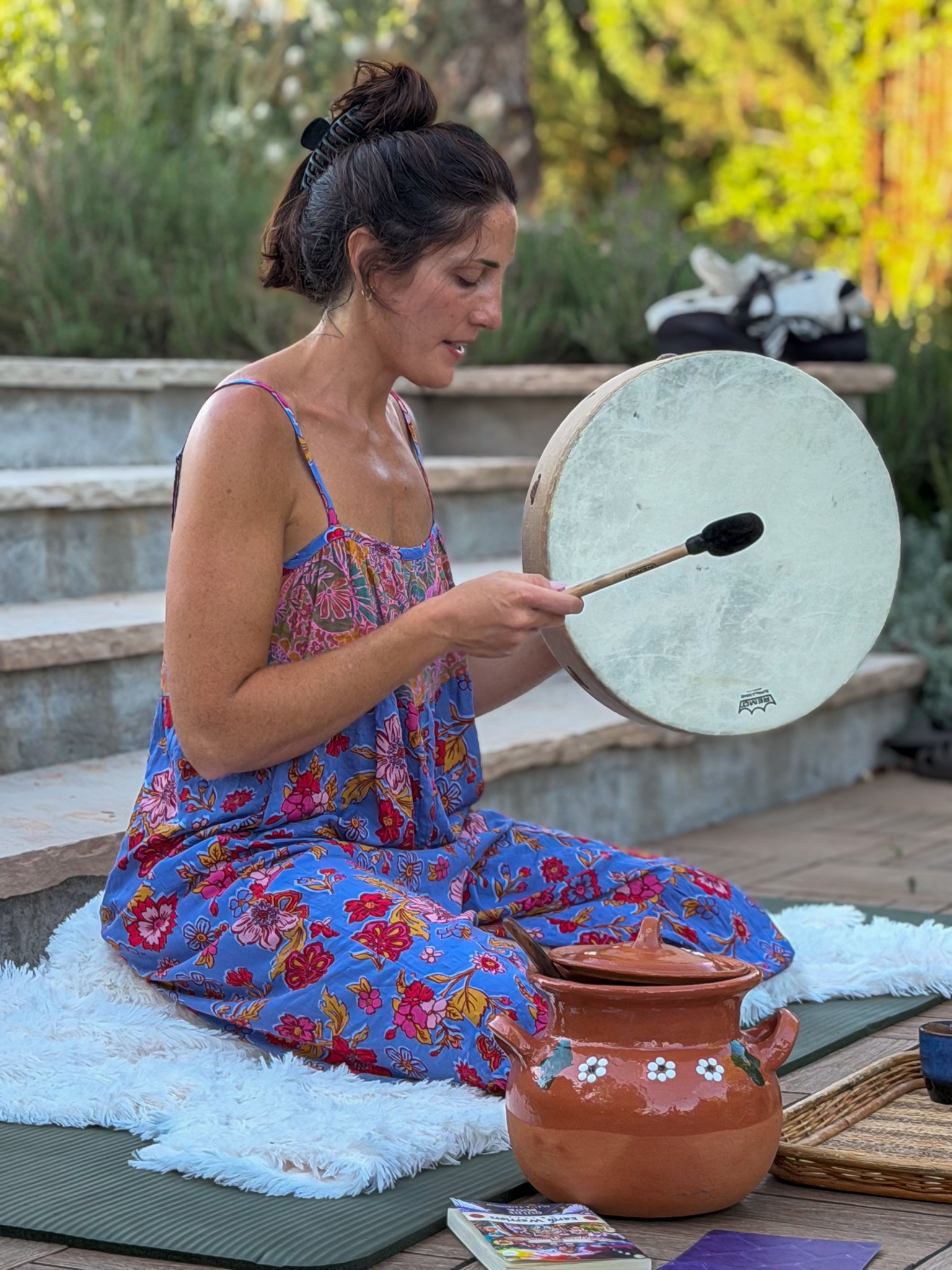A woman sitting with a drum in her hands outdoors, wearing a colorful floral dress, with a pottery vase and a magazine in front of her on a wooden surface, surrounded by greenery.