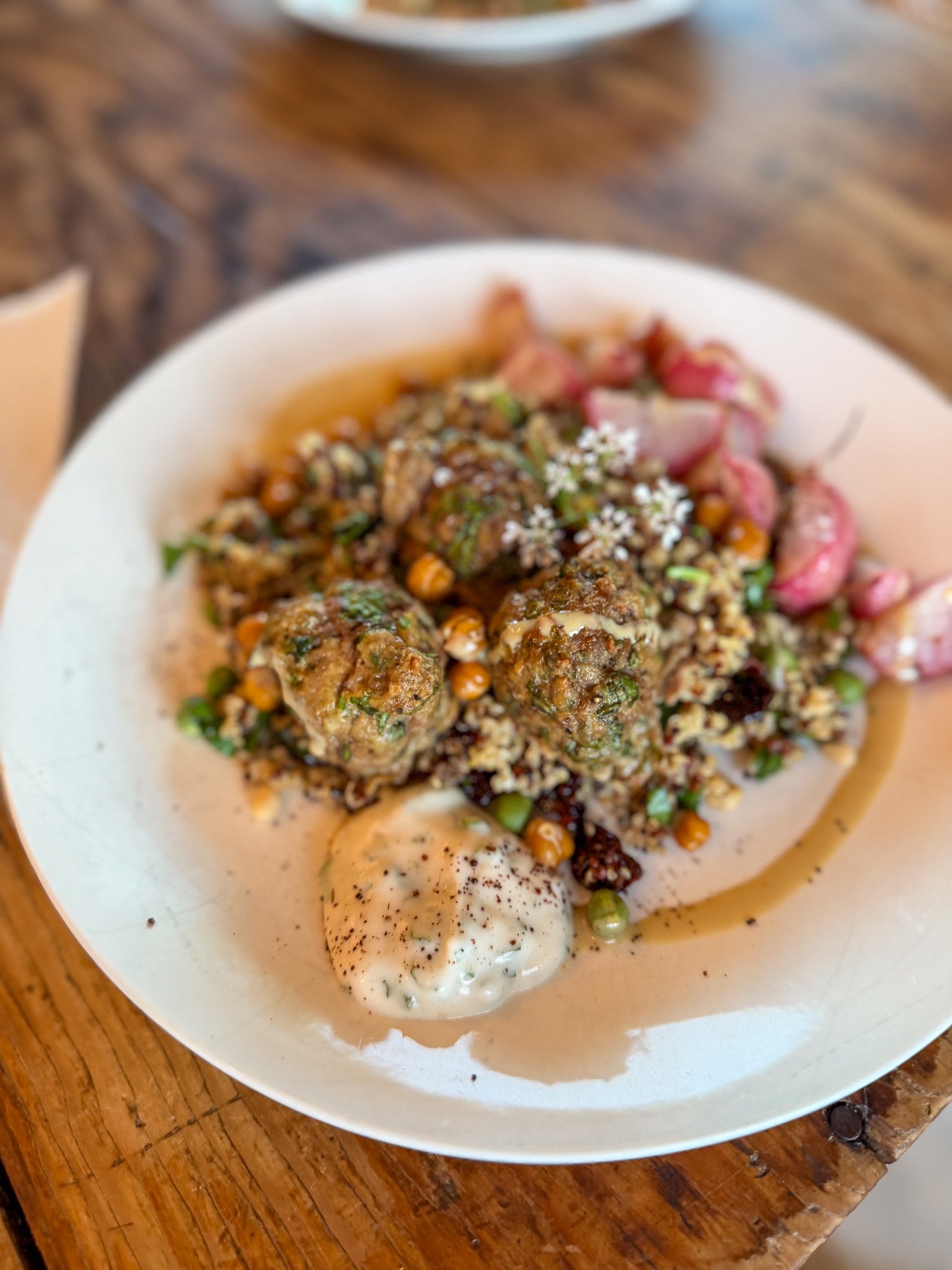 A white bowl contains an assortment of food items, including meatballs, couscous or quinoa salad, roasted vegetables, and a dollop of creamy sauce with herbs, all served on a wooden table.