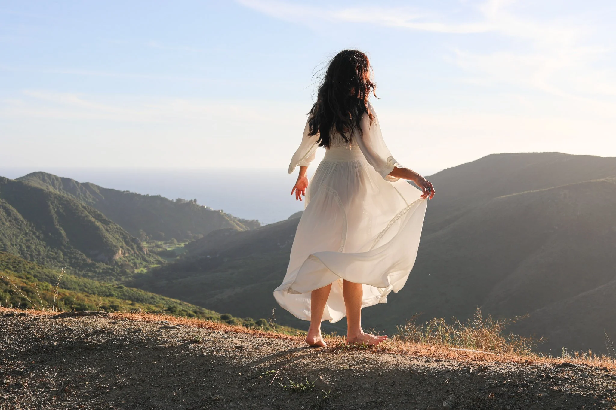A woman in a white dress walking barefoot on a mountain trail with scenic green hills and a distant ocean in the background during sunset or sunrise.