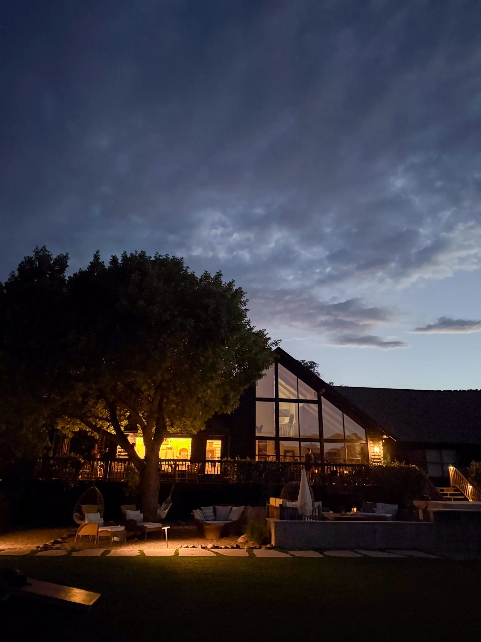 Nighttime scene of a backyard with a house that has large windows emitting warm light, several chairs, a table with an umbrella, trees, and a sky with clouds.