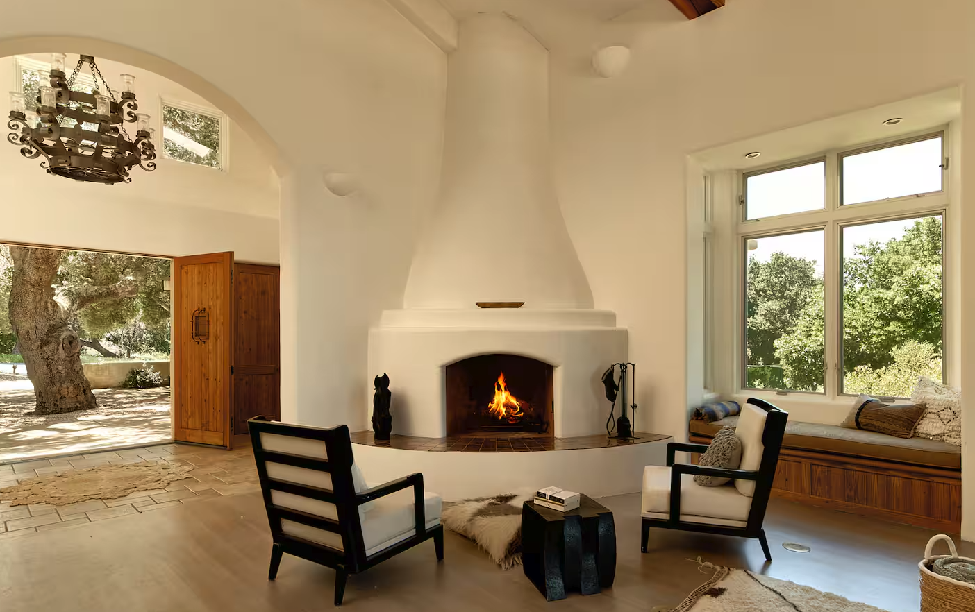 Living room with a white fireplace, two black and white armchairs, a window seat with cushions, a large window showing trees, and a door leading outside to a yard with a large tree.