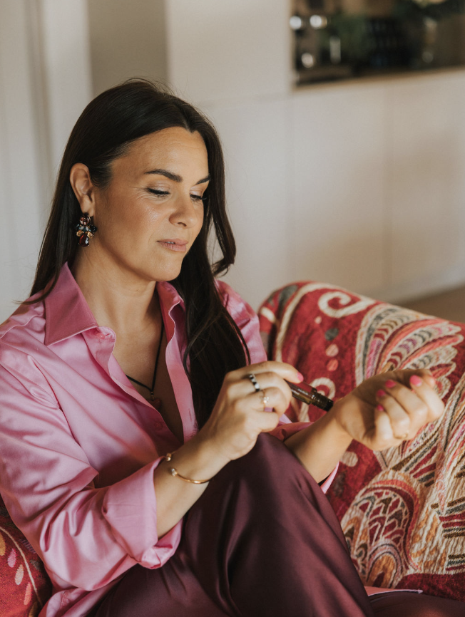 Femme assise sur un canapé, maquillant son ongle avec un vernis à ongles. Porte un chemisier rose et des boucles d'oreilles colorées.
