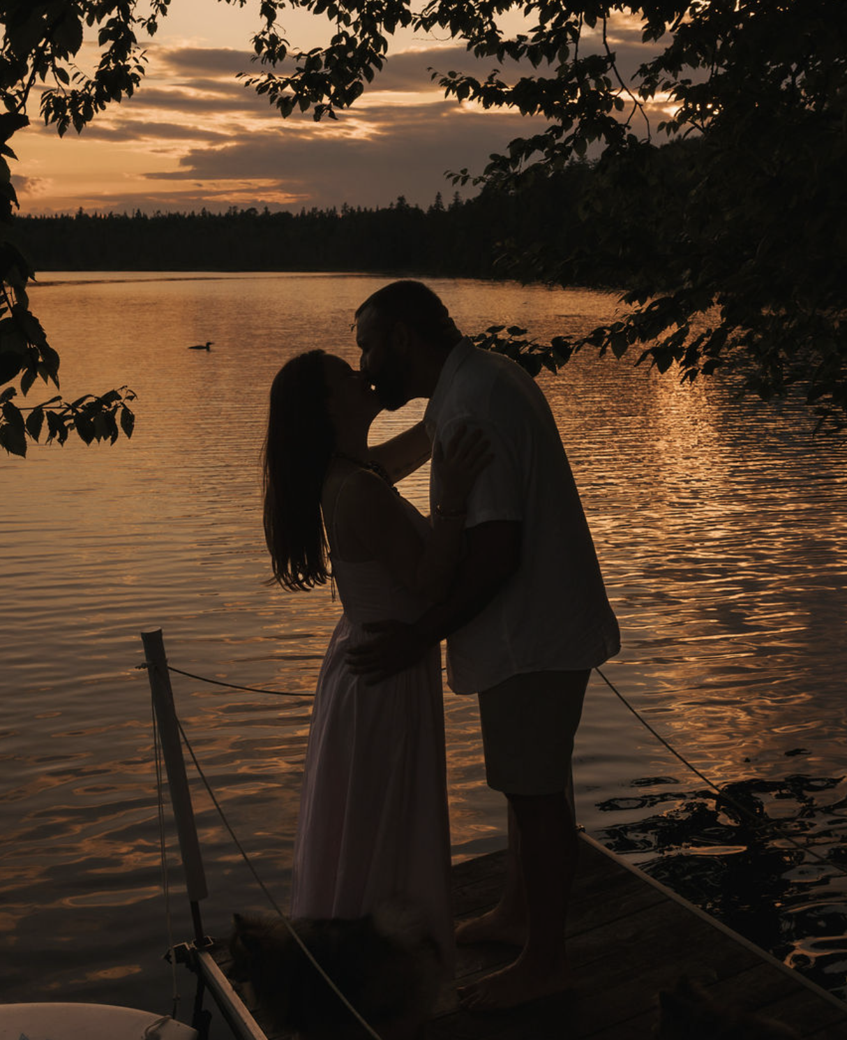 Un couple s'embrasse sur un pont au coucher du soleil, avec un lac en arrière-plan et des arbres encadrant la scène.