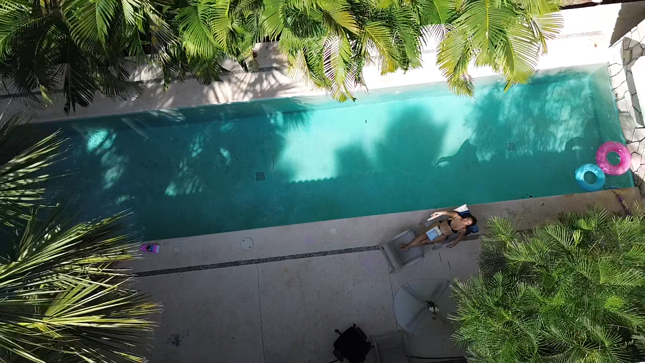 Vue aérienne d'une piscine avec une femme assise sur un fauteuil sur la terrasse, entourée de palmiers.