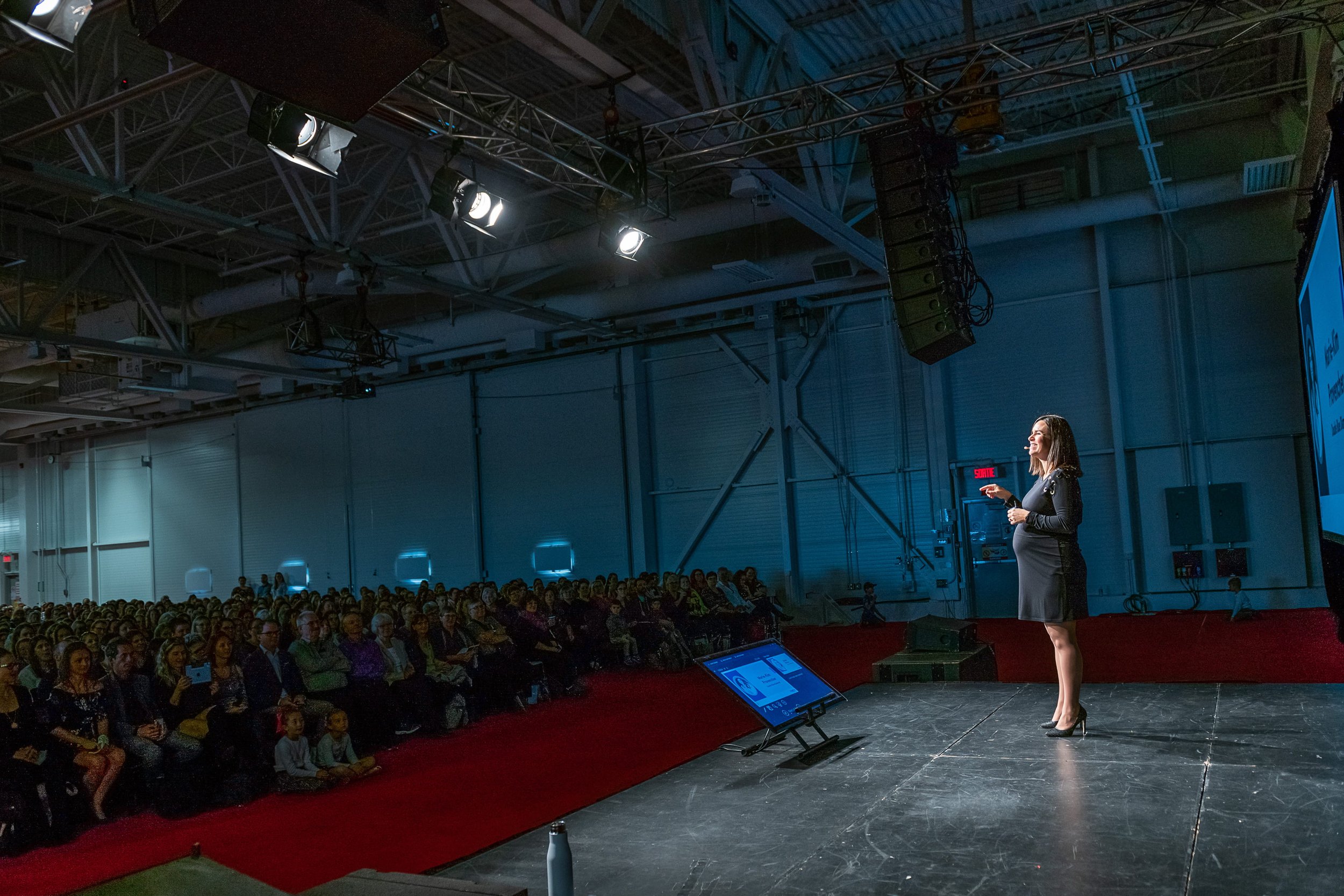 Une femme parle en public lors d'une conférence, face à une grande audience dans une salle avec éclairage professionnel.