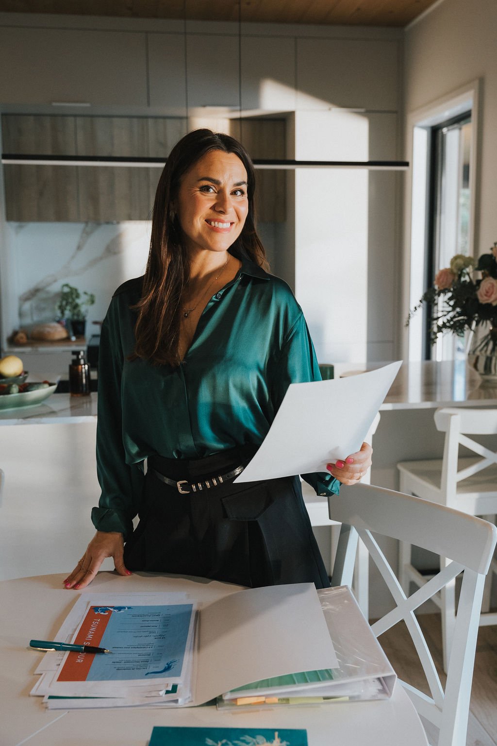 Femme souriante en tenue professionnelle, tenant des documents, dans une cuisine lumineuse avec table, chaises et décor floral.