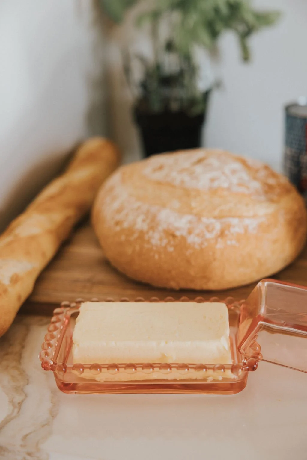 Pain frais, beurre dans un plat en verre, baguette de pain, pain rond, plante verte en arrière-plan.