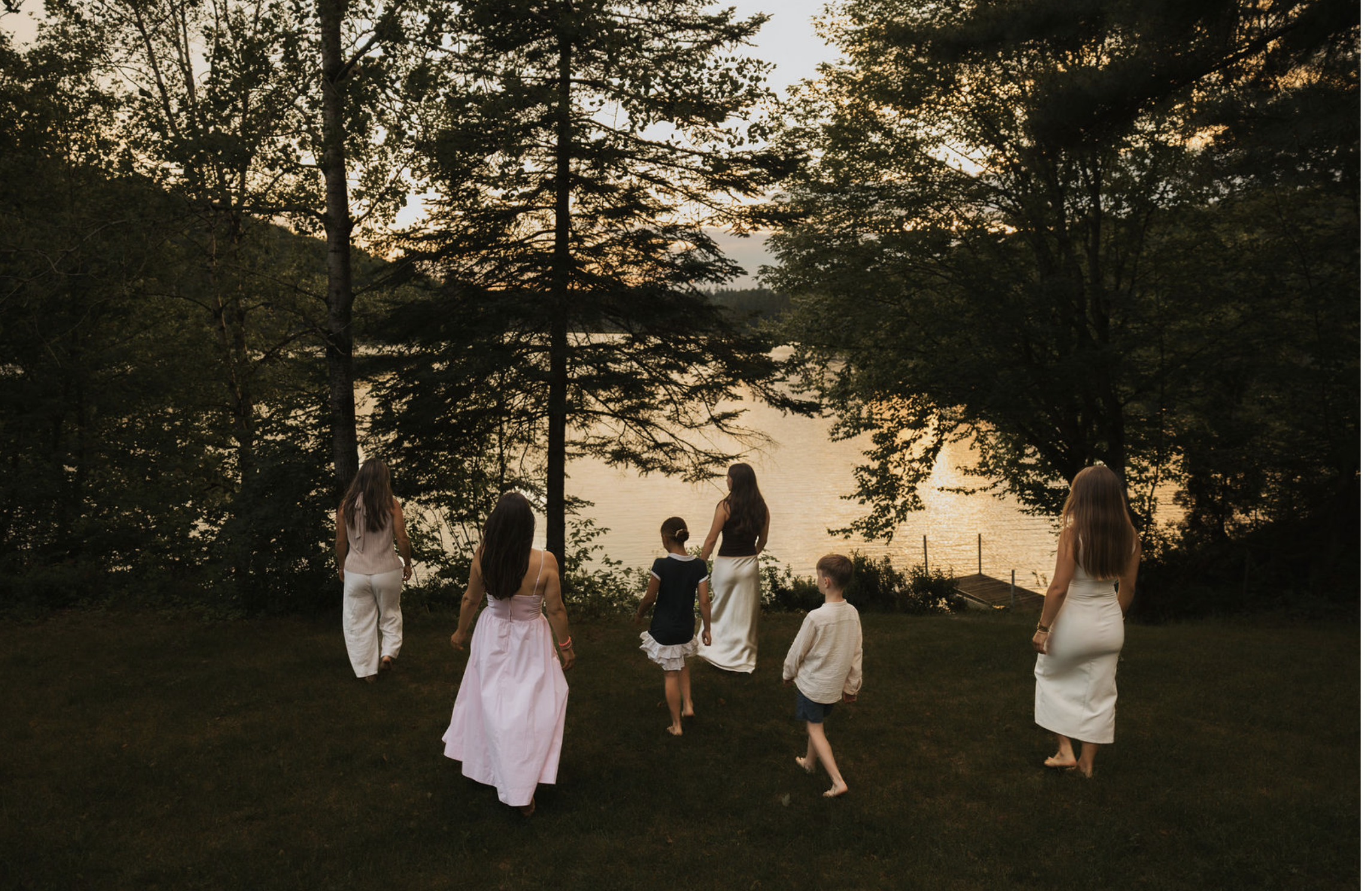 Groupe de personnes, principalement des femmes et des enfants, marchant dans une forêt au coucher du soleil près d'un lac. La scène dégage une atmosphère paisible et naturelle.