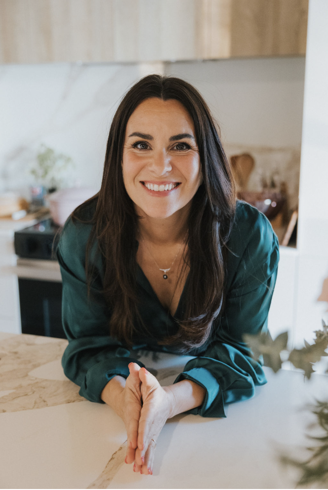 Femme souriante avec des cheveux bruns longs, portant une chemise verte, dans une cuisine moderne.