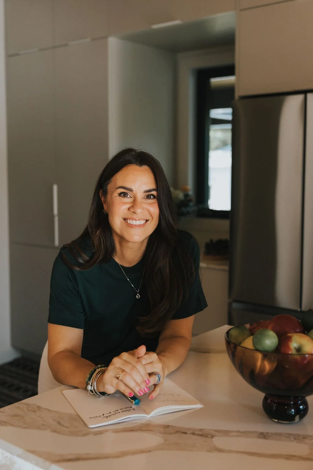 Une femme souriante avec des cheveux longs noirs, portant un t-shirt vert foncé, assise à une table en marbre dans une cuisine moderne, avec un bol de fruits et un carnet de notes devant elle.