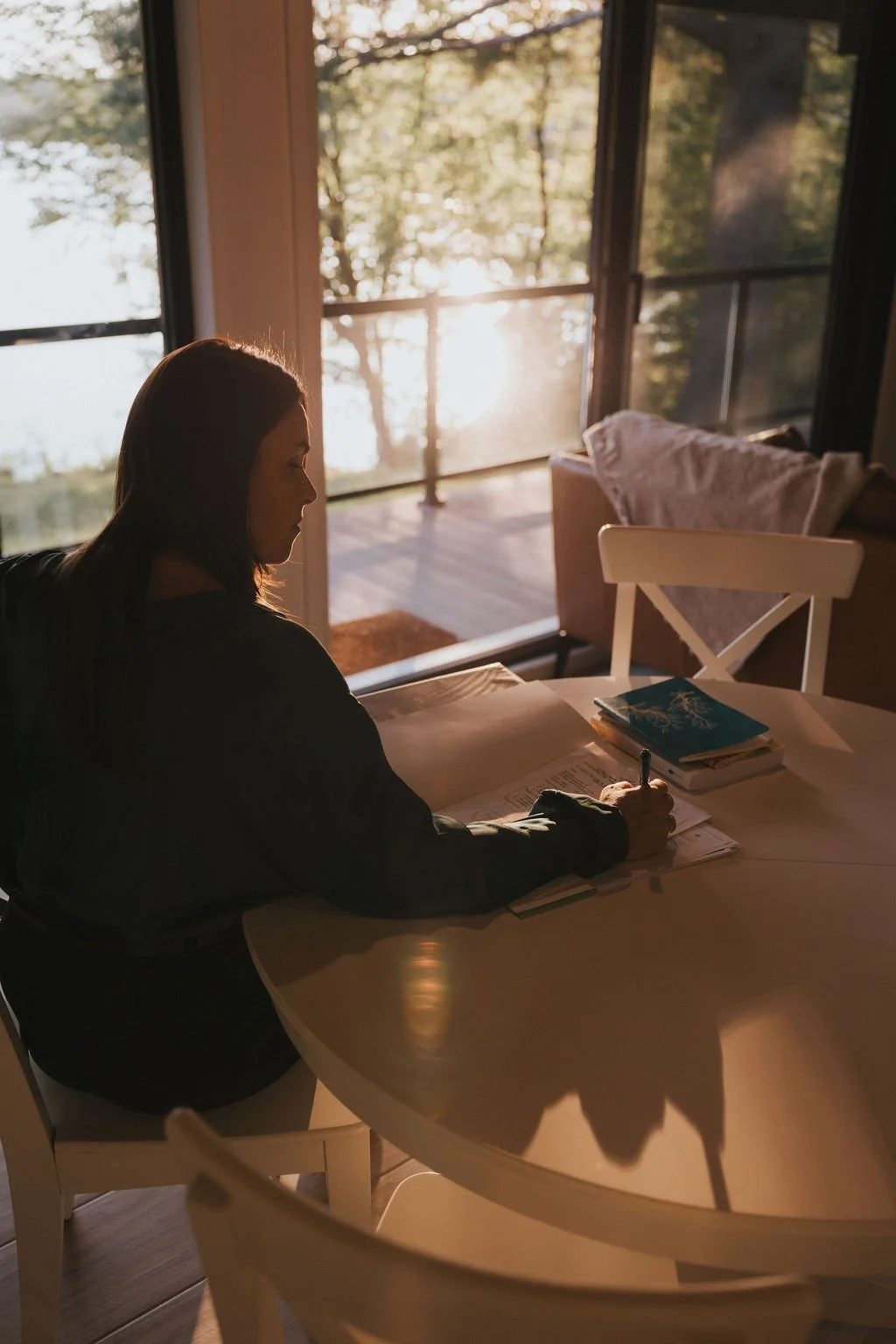 Femme assise à une table, écrivant, avec une vue sur la nature au coucher du soleil à l'arrière-plan.