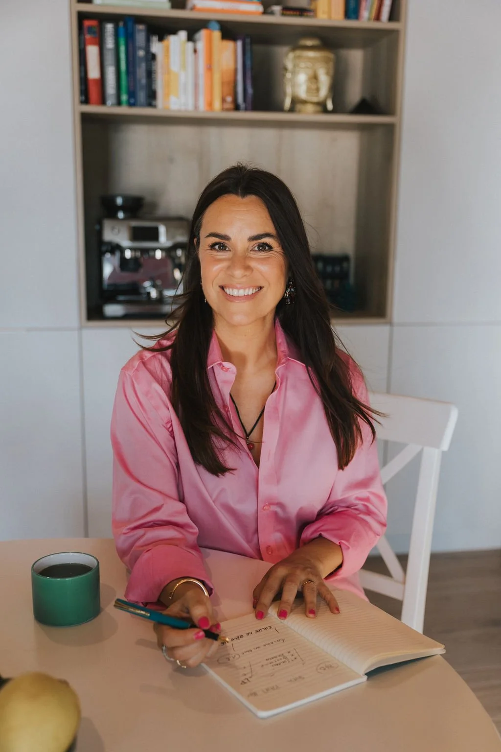 Une femme souriante assise à une table, portant une chemise rose,tenant un stylo et écrivant dans un carnet, avec une tasse de café à côté, dans un intérieur moderne.