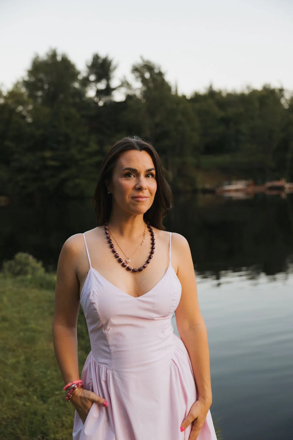 Femme portant une robe rose avec un collier en perles, se tenant près d'un lac au coucher du soleil.