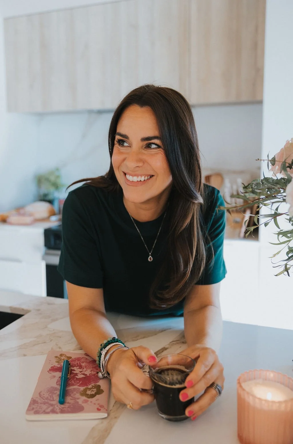 Une femme souriante à la maison, tenant une tasse de café ou de thé, avec un carnet et un stylo sur la table, décoration intérieure chaleureuse