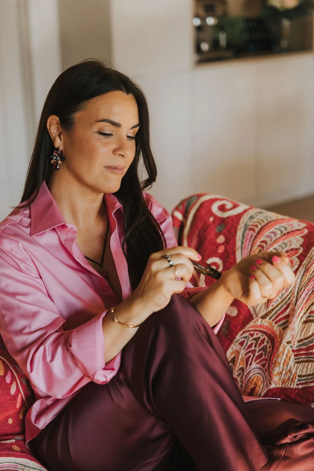 Une femme assise sur un canapé à motifs rouges, portant une chemise rose en satin et des boucles d'oreilles colorées, tient un roll-on et semble appliquer une lotion ou un parfum sur son poignet.