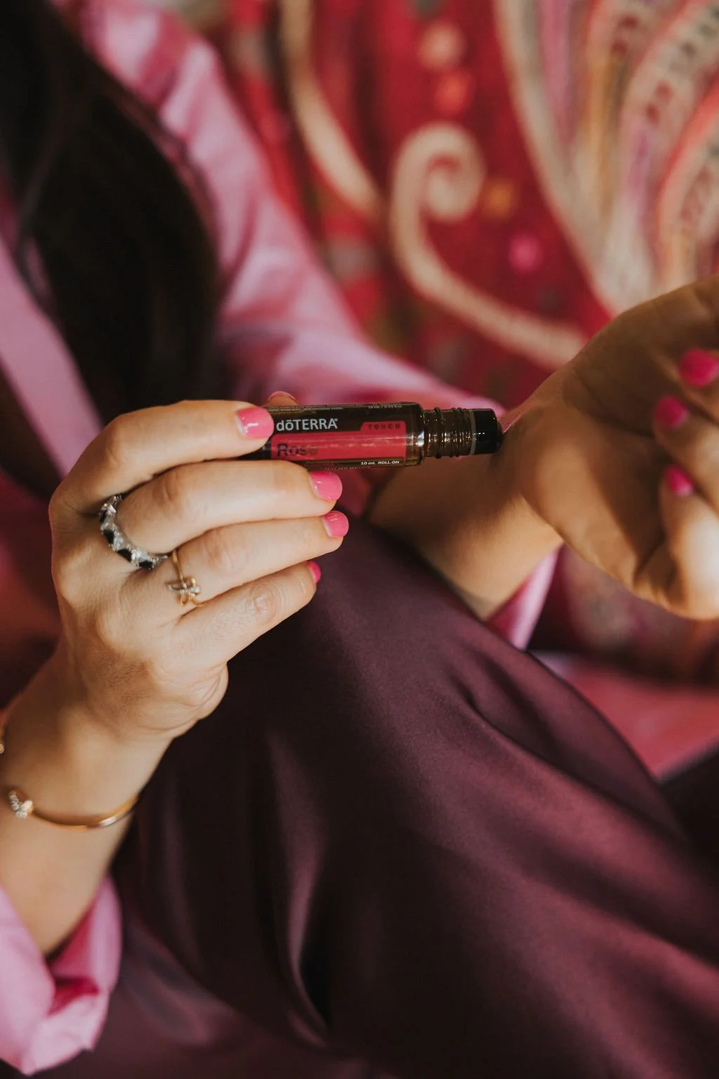 Main d'une personne tenant un rouleau d'huile essentielle doTERRA avec ongles and nails peint en rose, portant plusieurs bijoux, en train de s'appliquer de l'huile sur la peau. Personne vêtue de vêtements roses et mauves avec un motif floral.