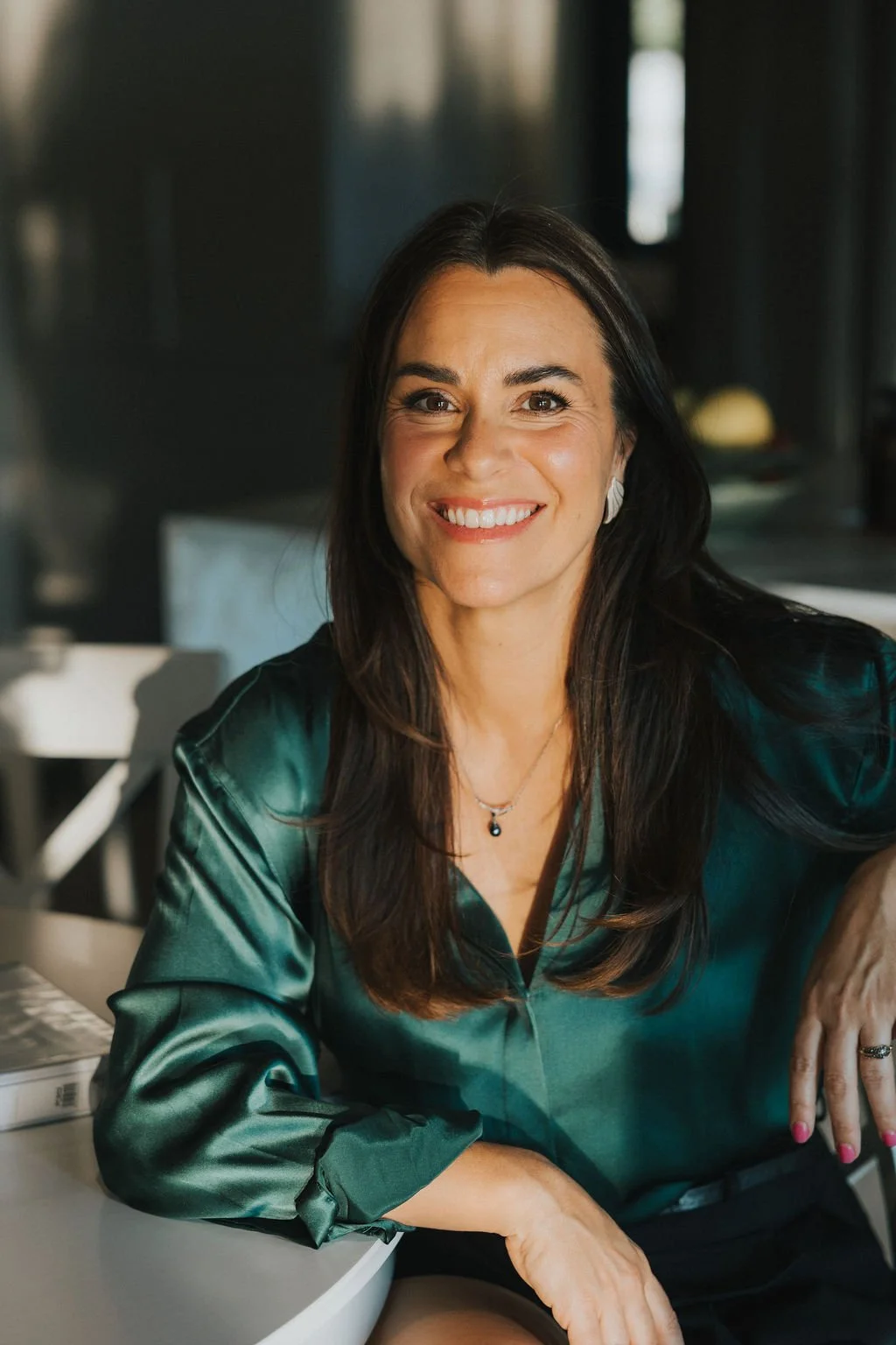 Une femme souriante aux cheveux bruns, portant un chemisier vert en satin, assise à un bureau.