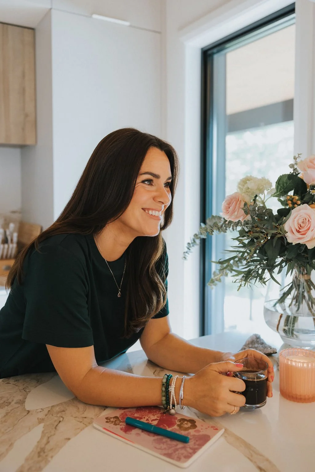 Une femme aux cheveux longs et bruns souriante, tenant une tasse de café ou de thé, assise à une table avec un carnet et un stylo, dans une cuisine lumineuse avec une grande fenêtre, un vase avec des roses