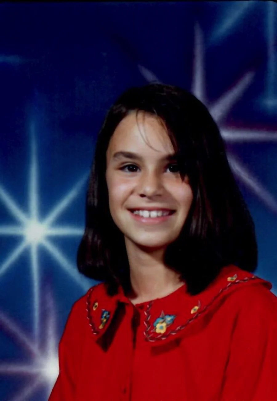 Une jeune fille souriante avec des cheveux noirs, portant une chemise rouge avec un col brodé de motifs colorés, pose devant un fond étoilé bleu.