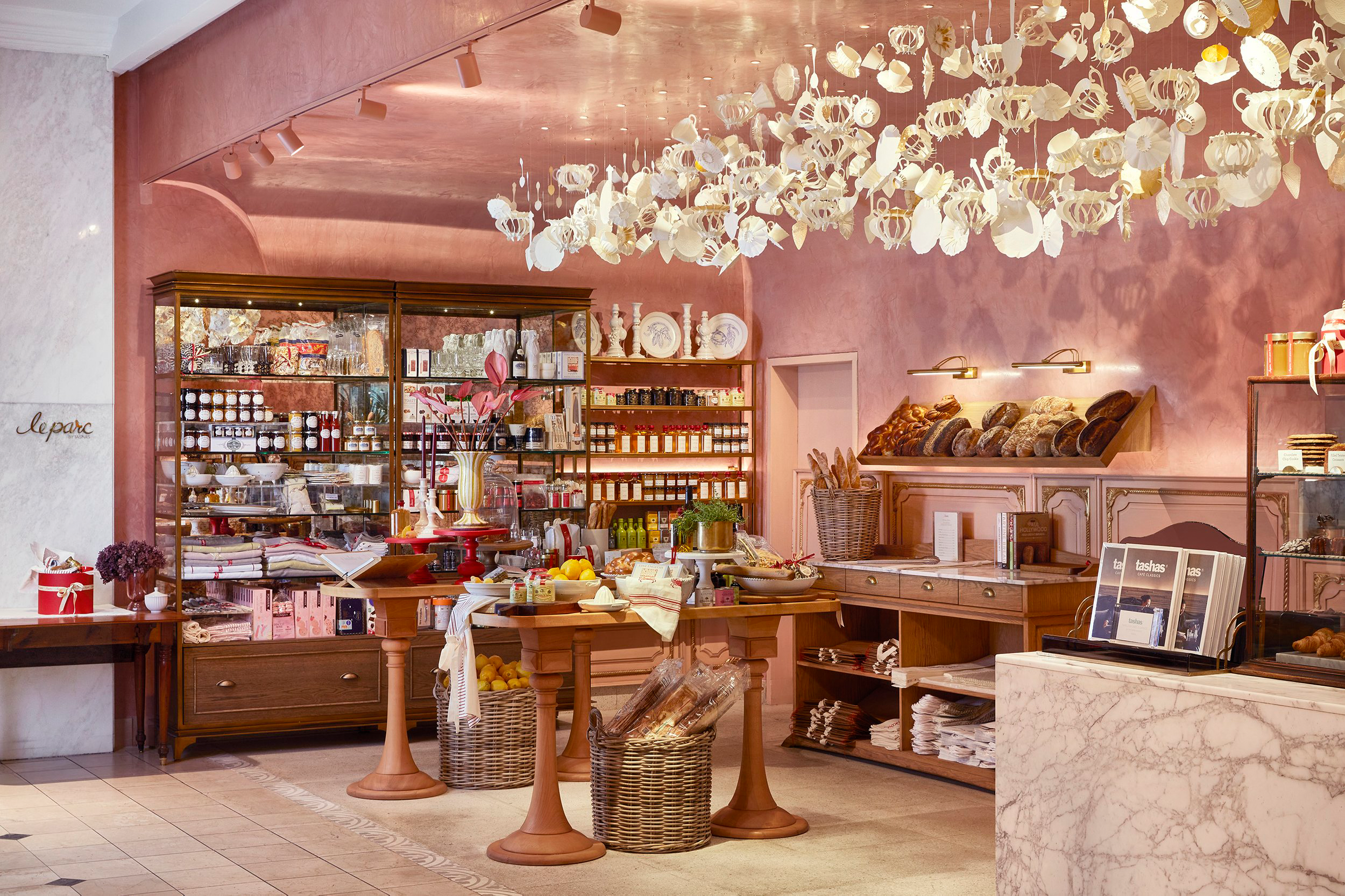 A cozy bakery or cafe interior with pink walls and wooden furniture. Display shelves are filled with various baked goods, jars, and decorative plates. A round table holds fresh fruit and baked items, while a basket contains baked bread. Overhead, whi
