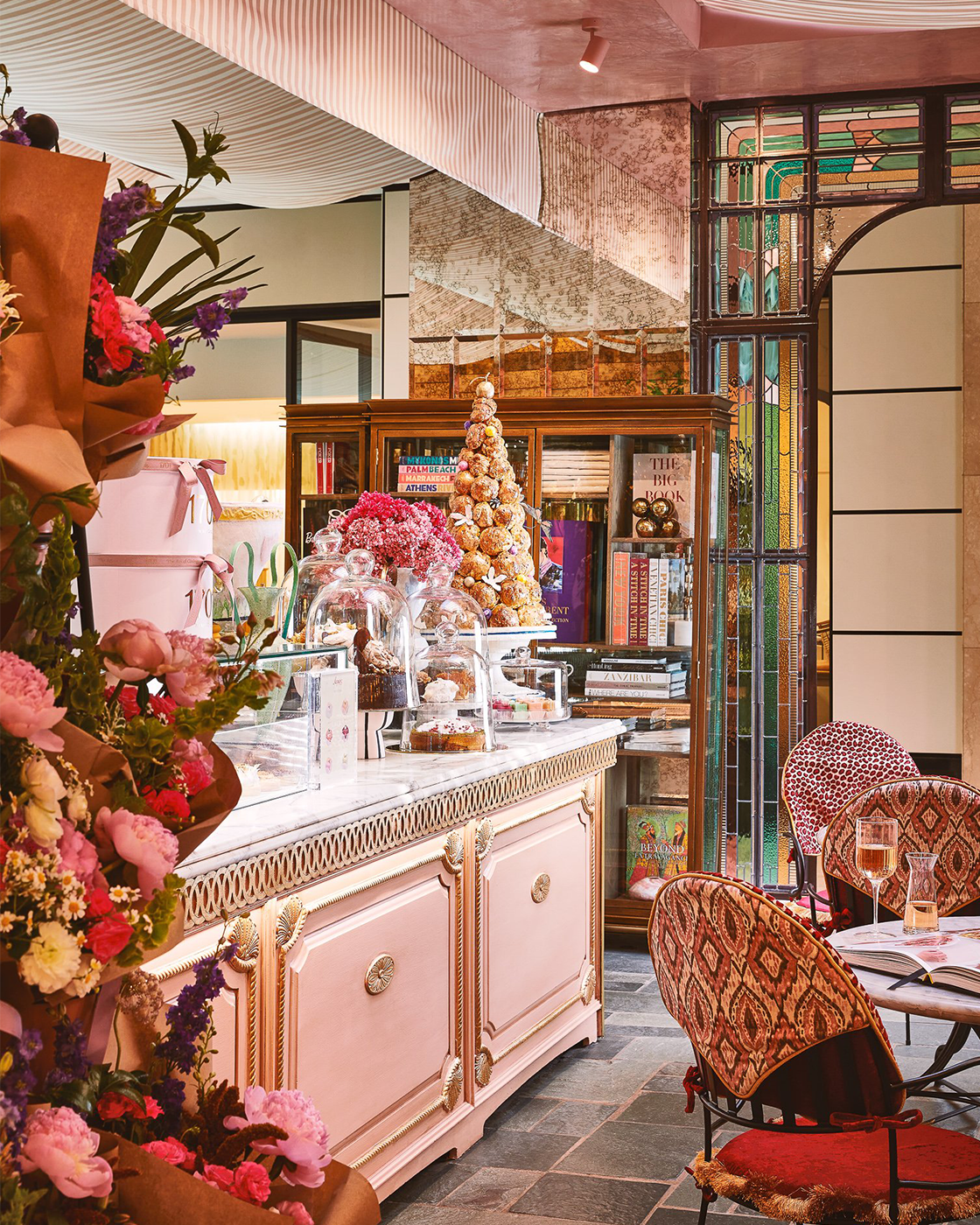 The interior of a decorative cafe or bakery featuring a pink, ornate buffet with glass display domes filled with pastries, flower arrangements, a small Christmas tree, and a cozy seating area with patterned chairs and framed stained glass windows.