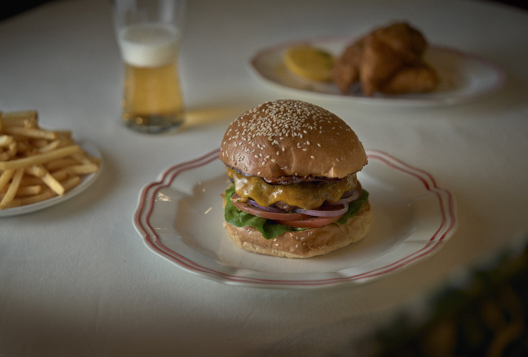 Close-up of a cheeseburger with lettuce, tomato, bacon, and cheese in a sesame seed bun on a decorative plate.