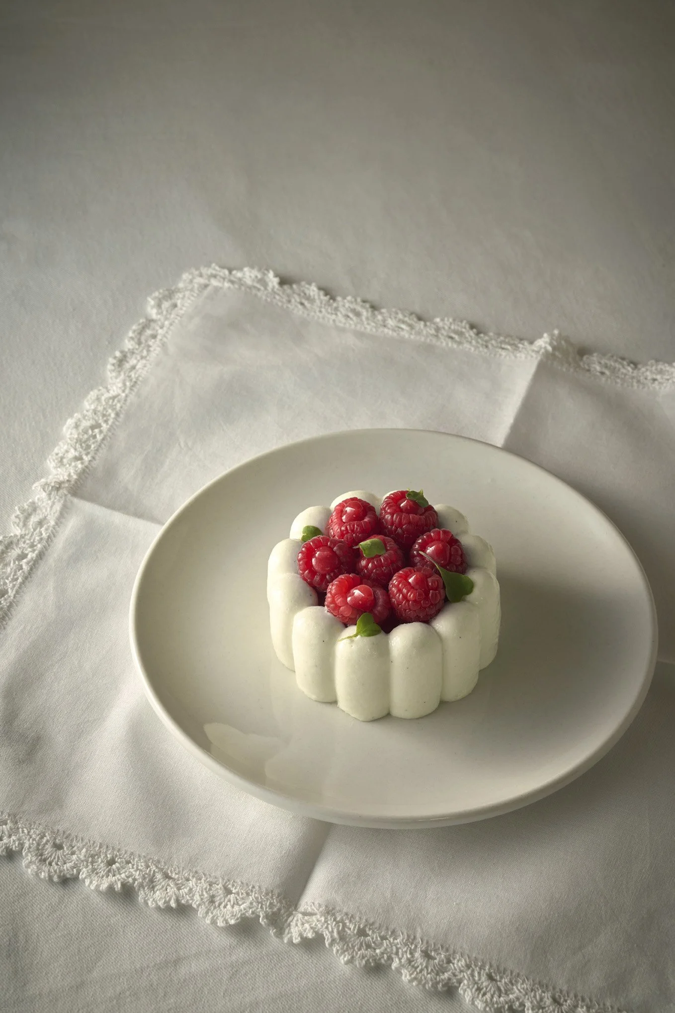 A small cake topped with fresh raspberries and white decorative dollops of cream, placed on a white plate on a white tablecloth with lace trim.
